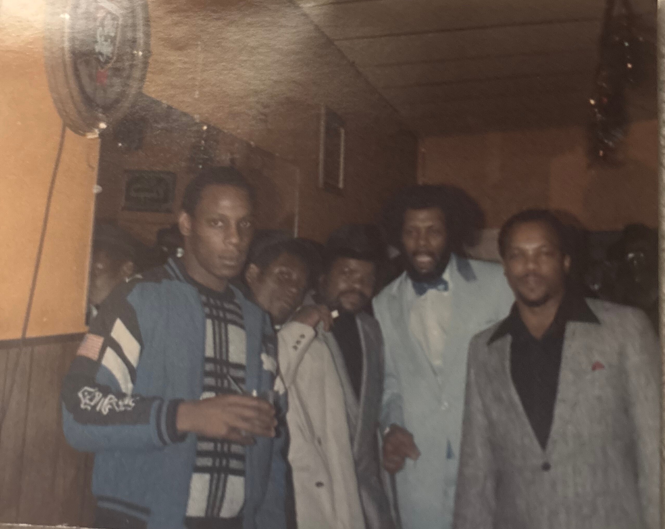 Group of men posing together indoors for a casual group photo in a dim room with wooden walls and warm lighting.