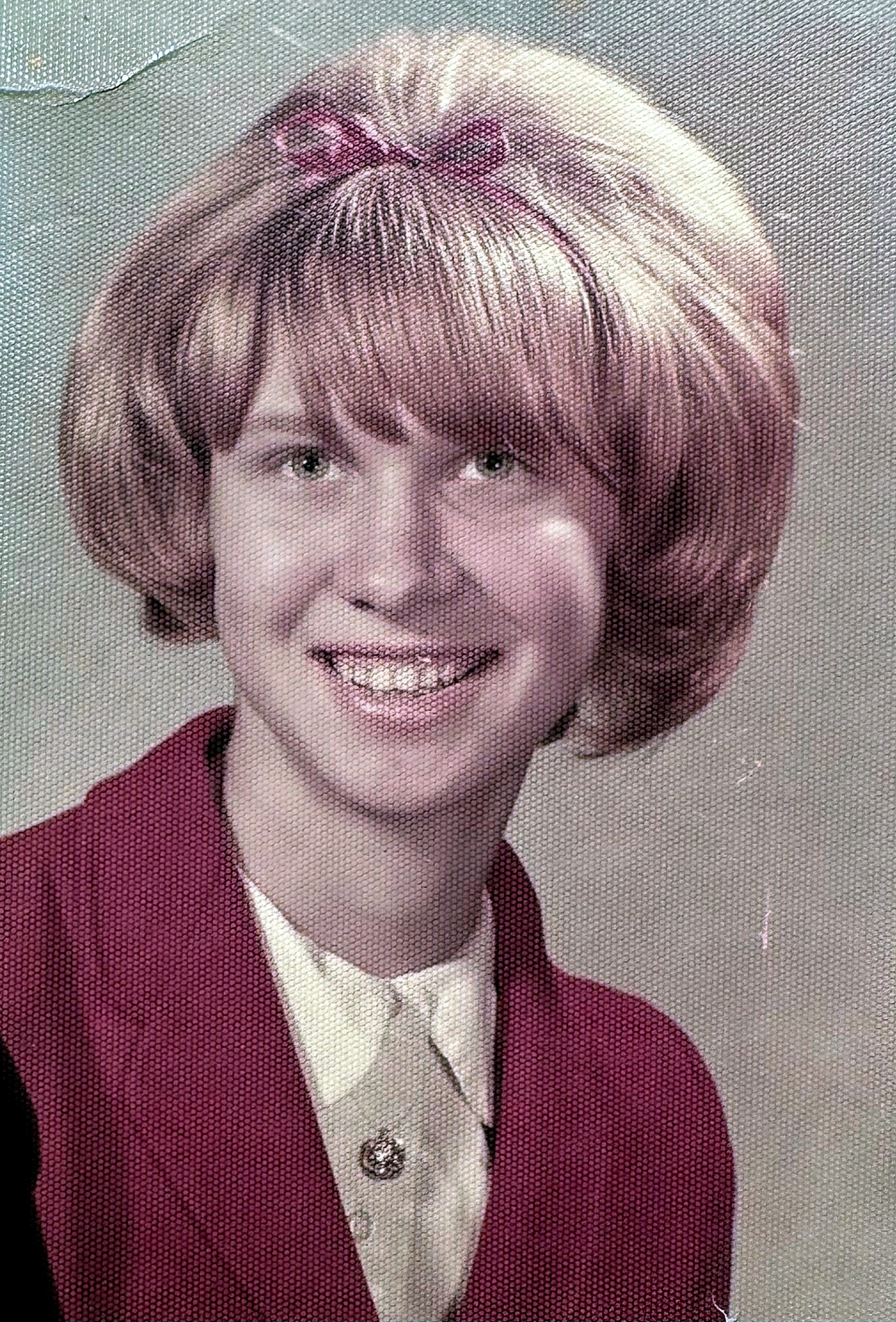 Smiling woman with short red hair wearing a pink blazer, white blouse, and a pin at the lapel in a studio portrait