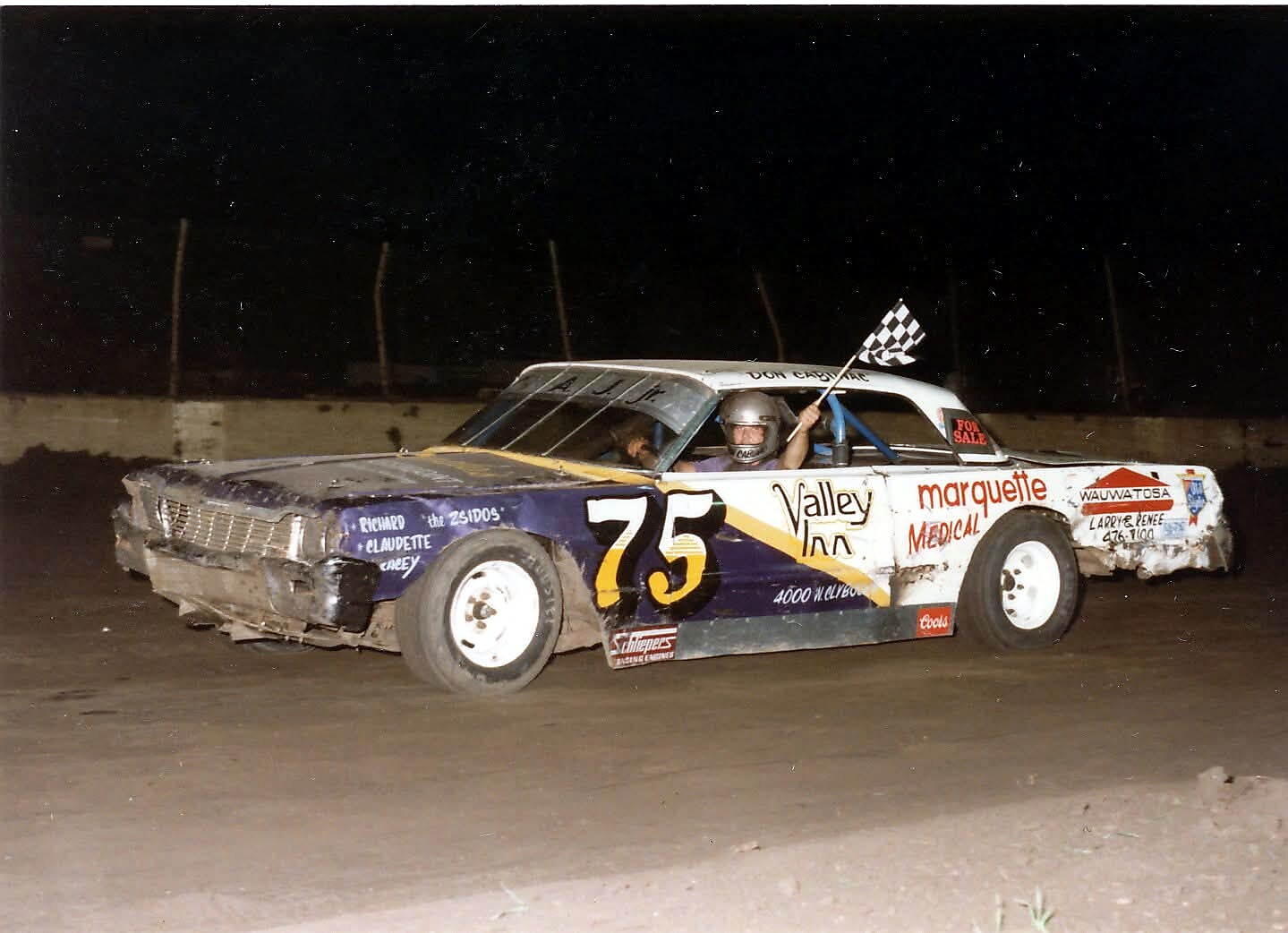 Nighttime dirt-track race car #75, driver in helmet waving a checkered flag from the window.