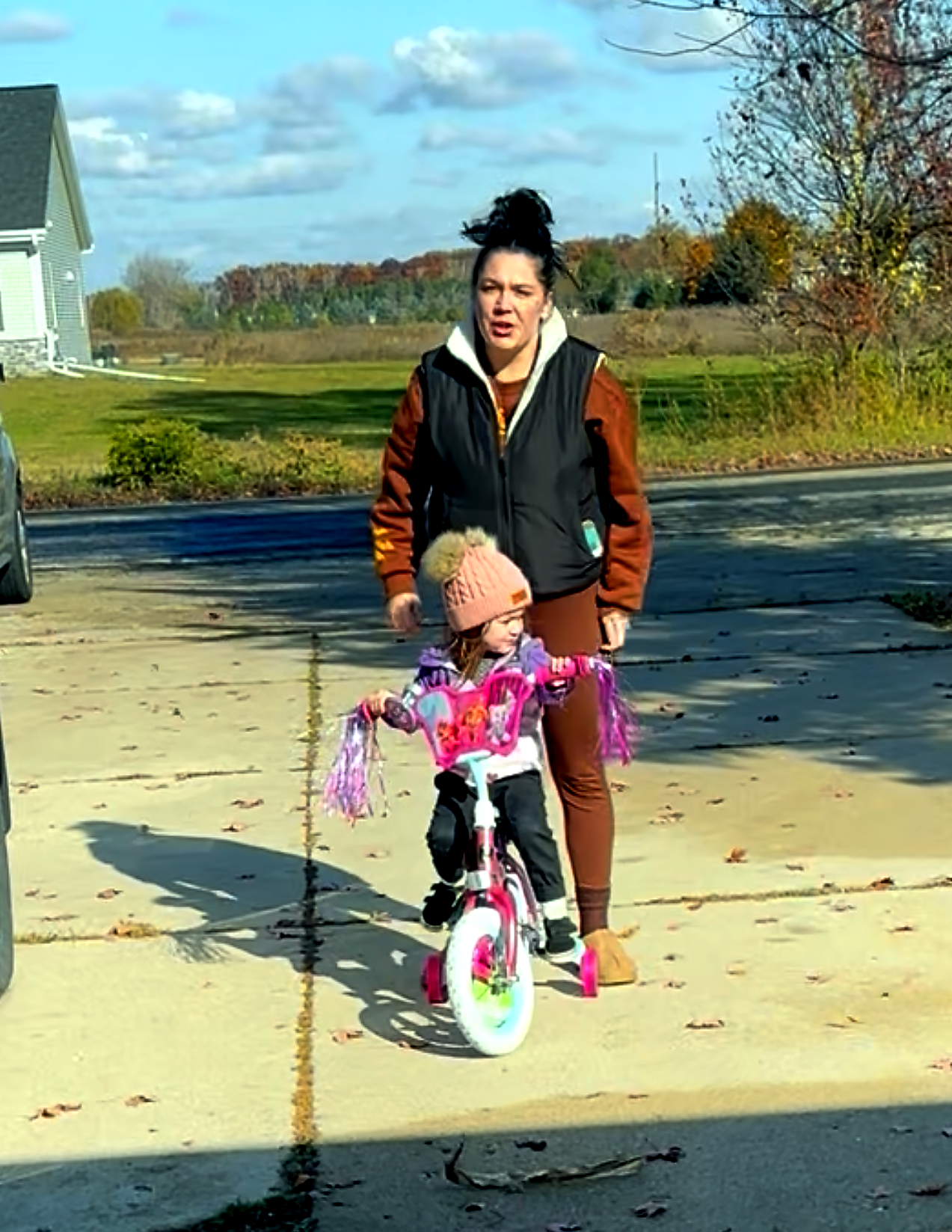 Woman helping a young girl ride a pink training-wheel bike outdoors on a sunny autumn day.