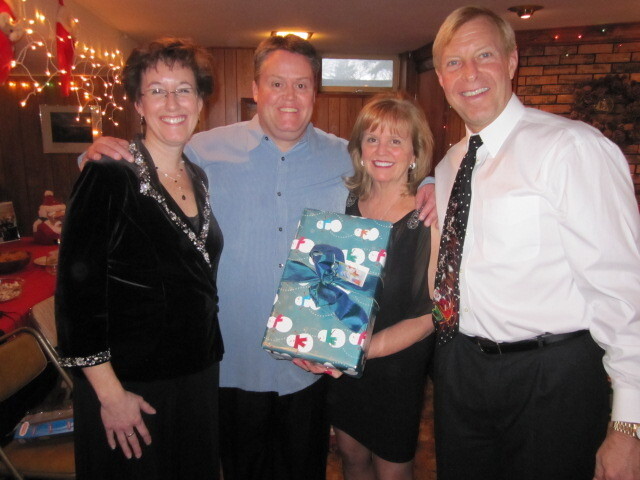 Four adults posing together at a holiday party, smiling; a wrapped gift is held by the woman second from the right, with festive lights and a decorated room in the background.