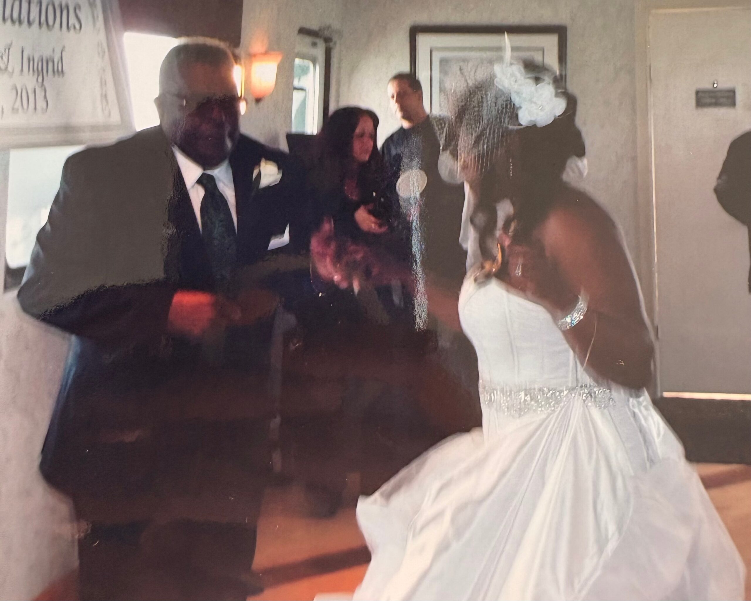 Bride in a white dress standing with guests in a dim room; a ‘congratulations’ banner partially visible on the left.