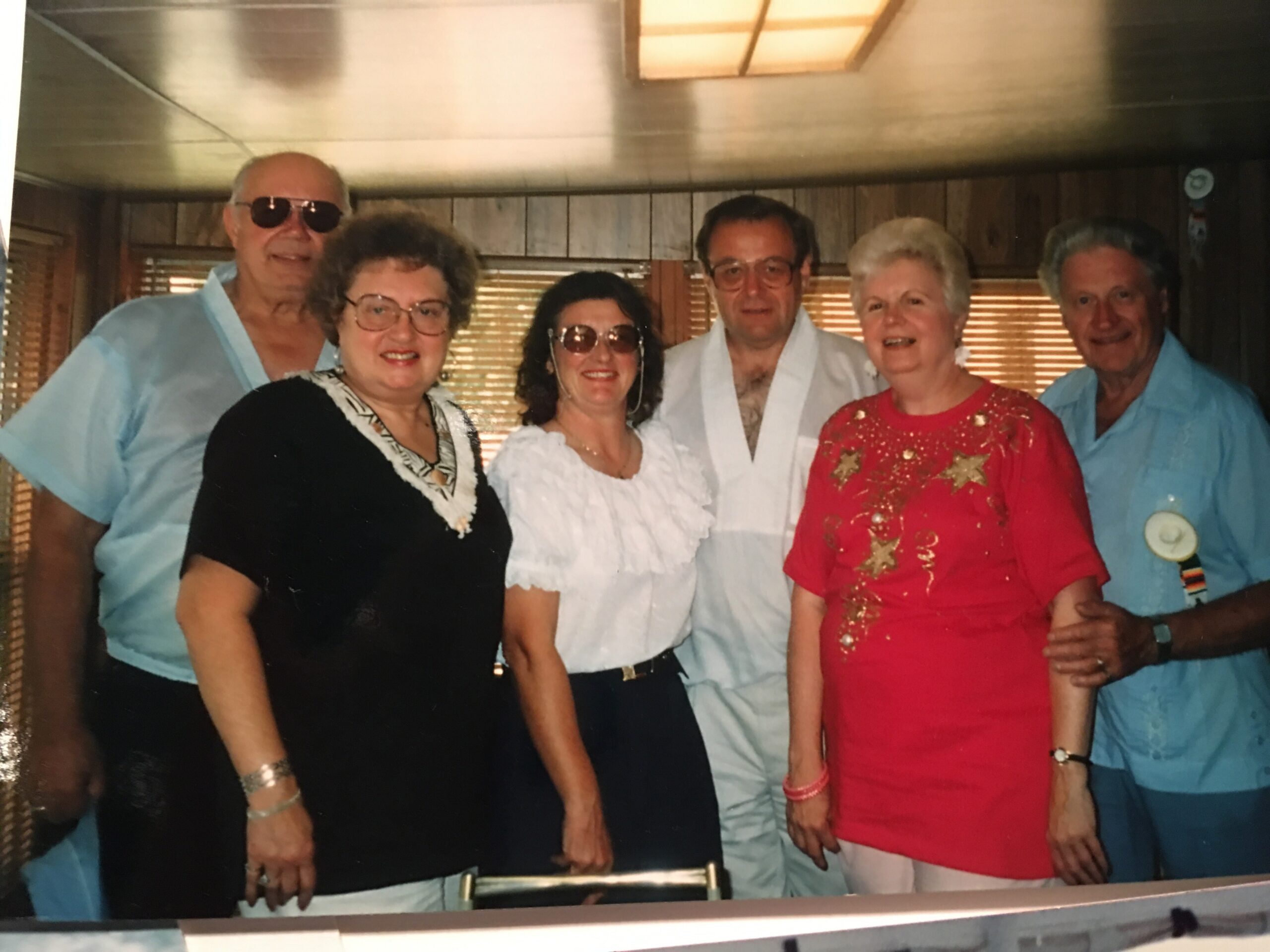 Six adults posing for a photo indoors in colorful retro outfits, smiling at the camera in a wood-paneled room.