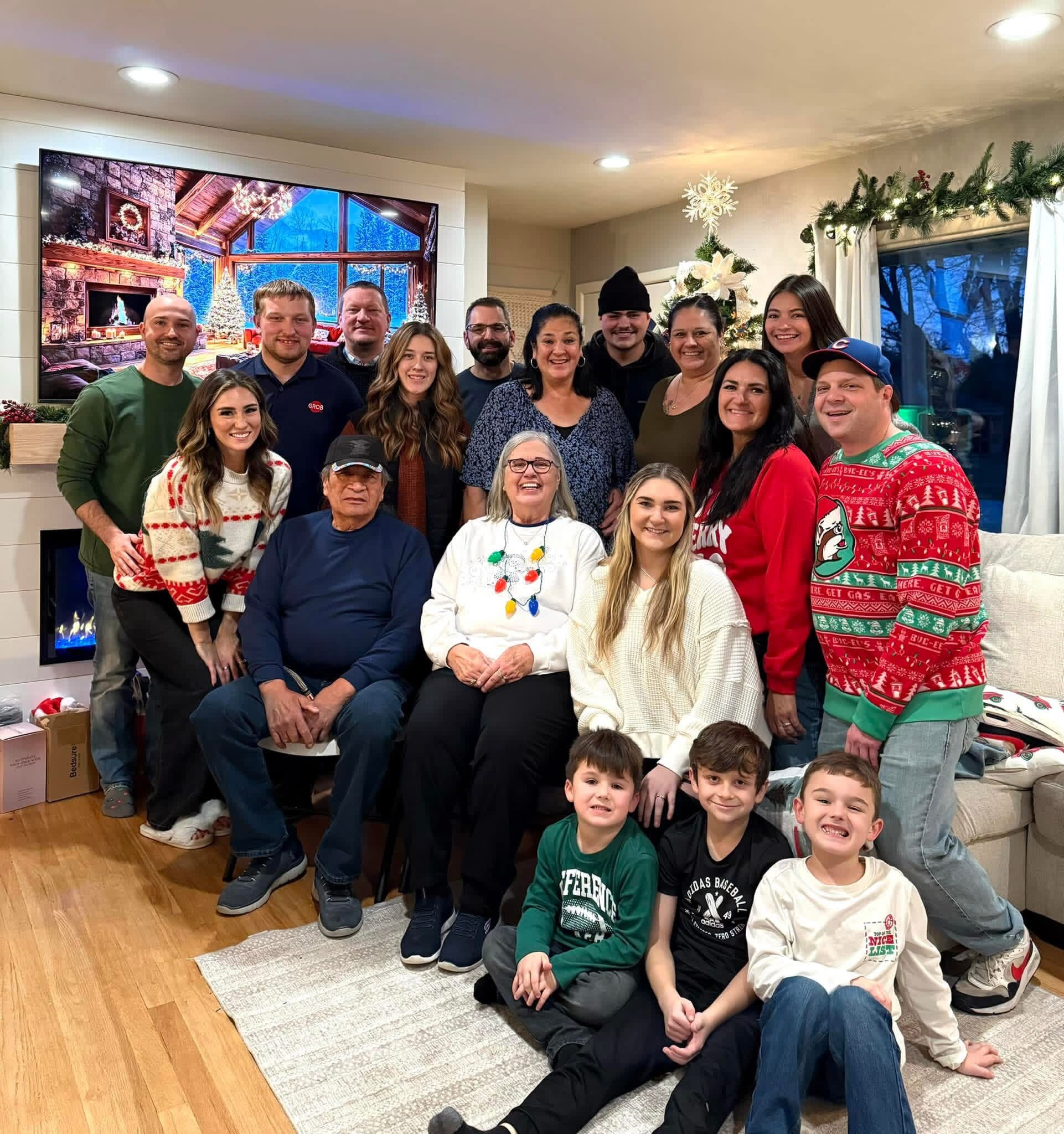 Large group of adults and children posing for a holiday photo indoors, with Christmas tree and festive decorations in the background.