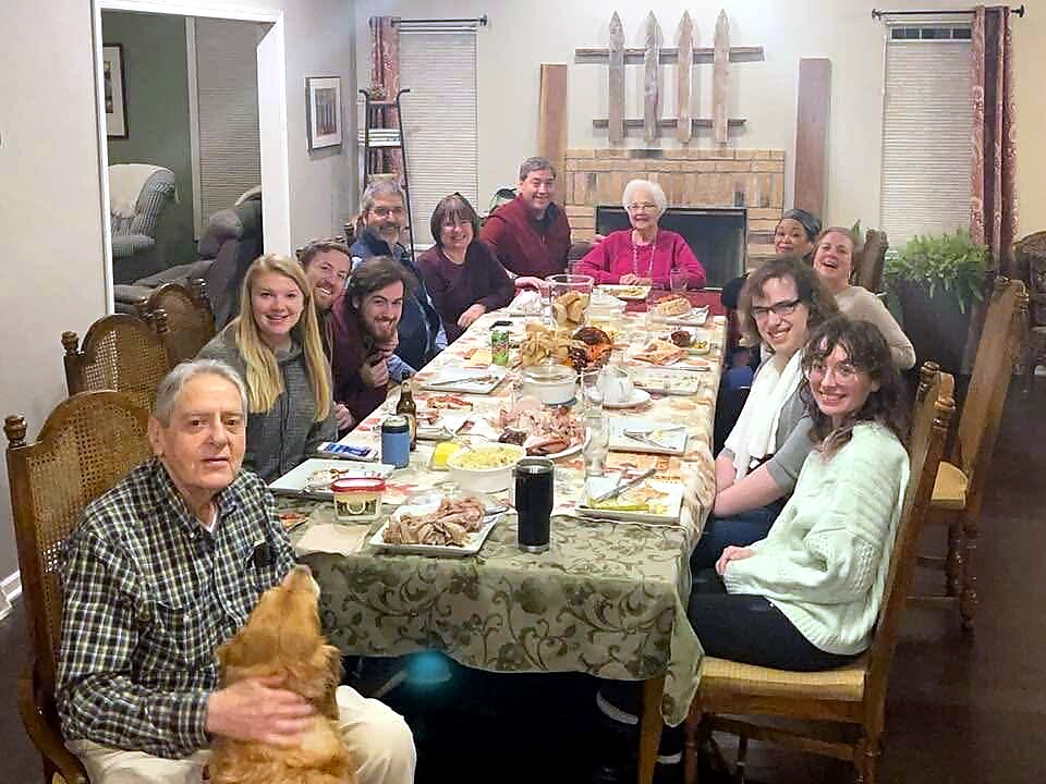 Family gathered around a long dining table-sharing a holiday meal in a cozy living room with a fireplace in the background, everyone smiling at the camera.