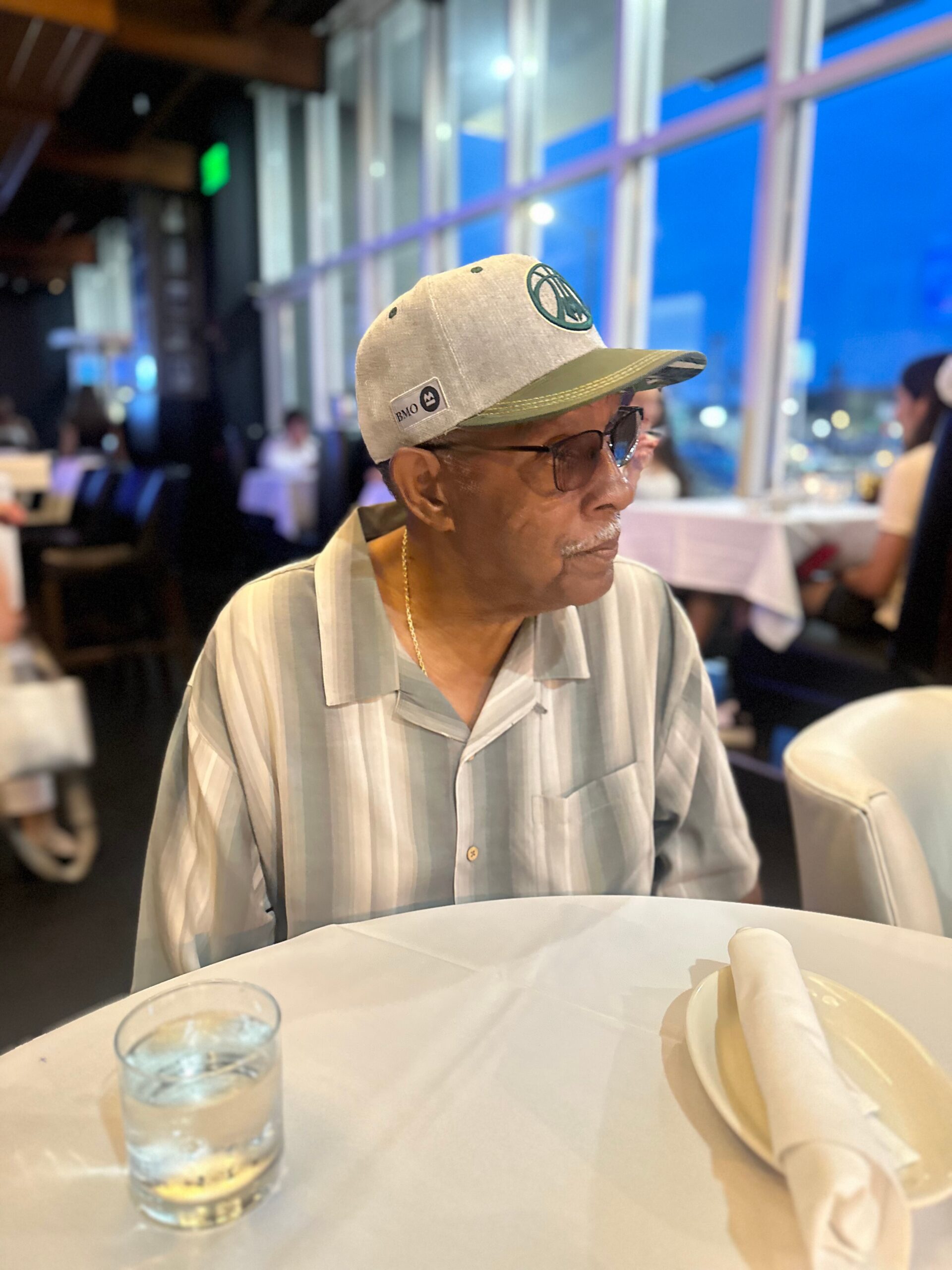Older man wearing a striped button-up shirt and cap sits at a dining table in a restaurant, glasses on, with a water glass and utensils in the foreground.