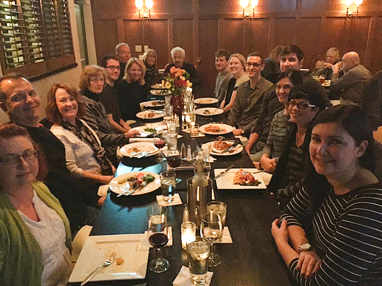 Group of adults seated around a long dinner table in a warmly lit restaurant, with plated meals, drinks, and a flower centerpiece.