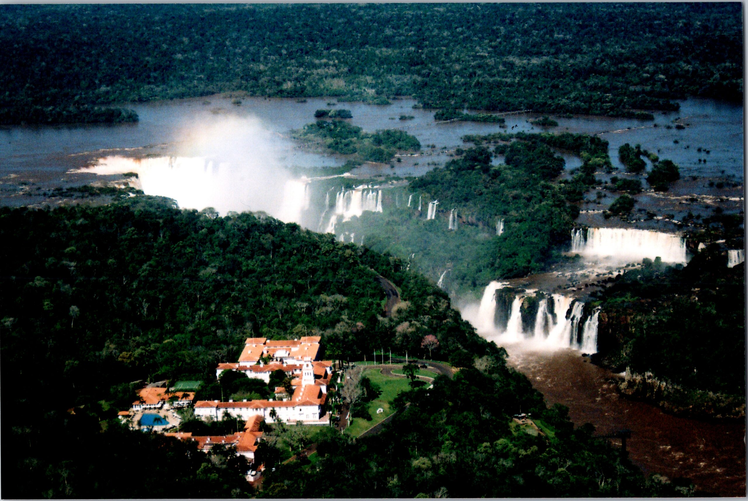 Aerial view of Iguazú Falls with multiple cascades surrounded by dense rainforest and a red-roofed resort in the foreground.