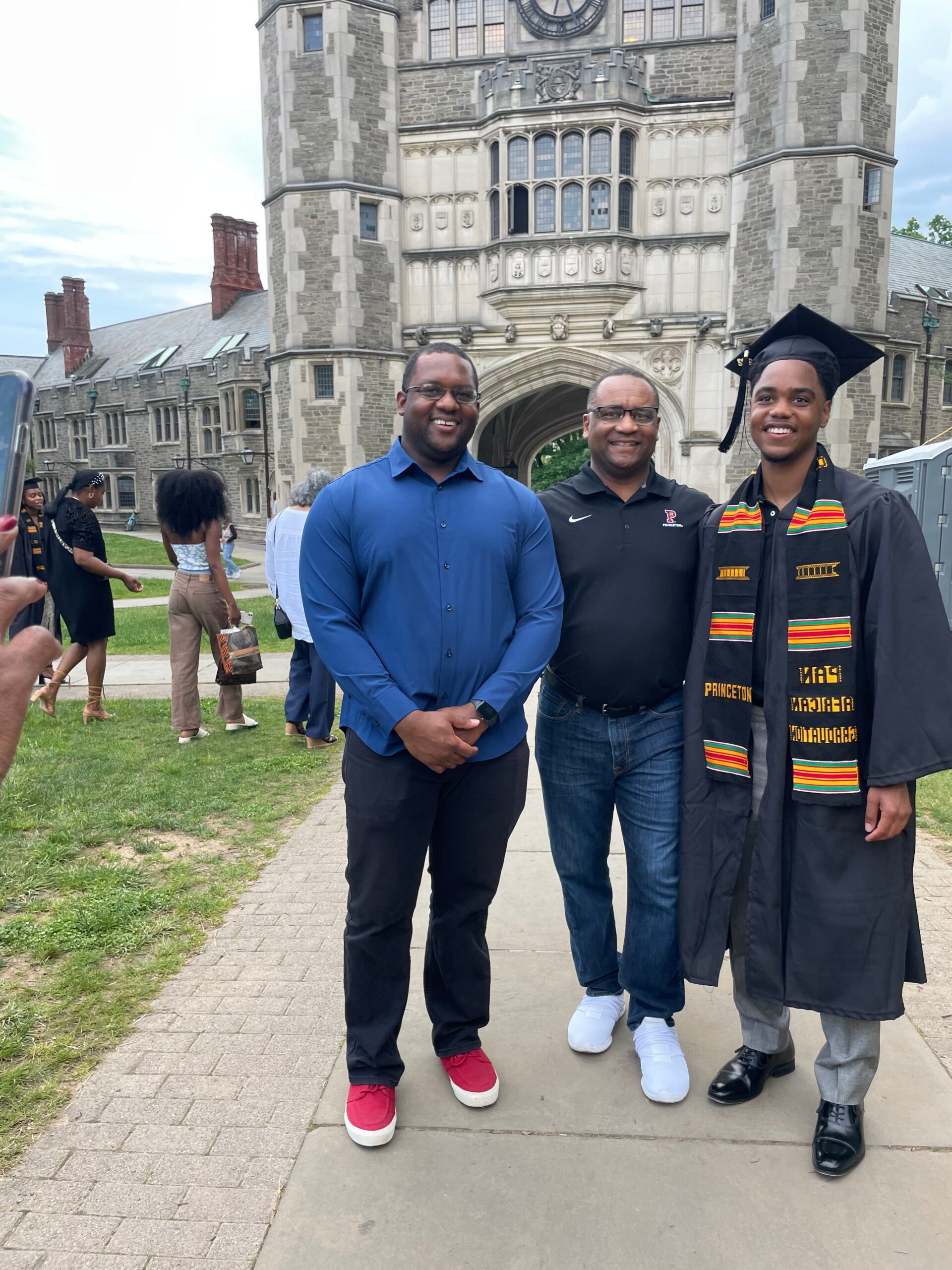 Three men posing for a graduation photo outside a historic campus building with a large archway; one graduate in cap and gown with a colorful stole stands on the right.