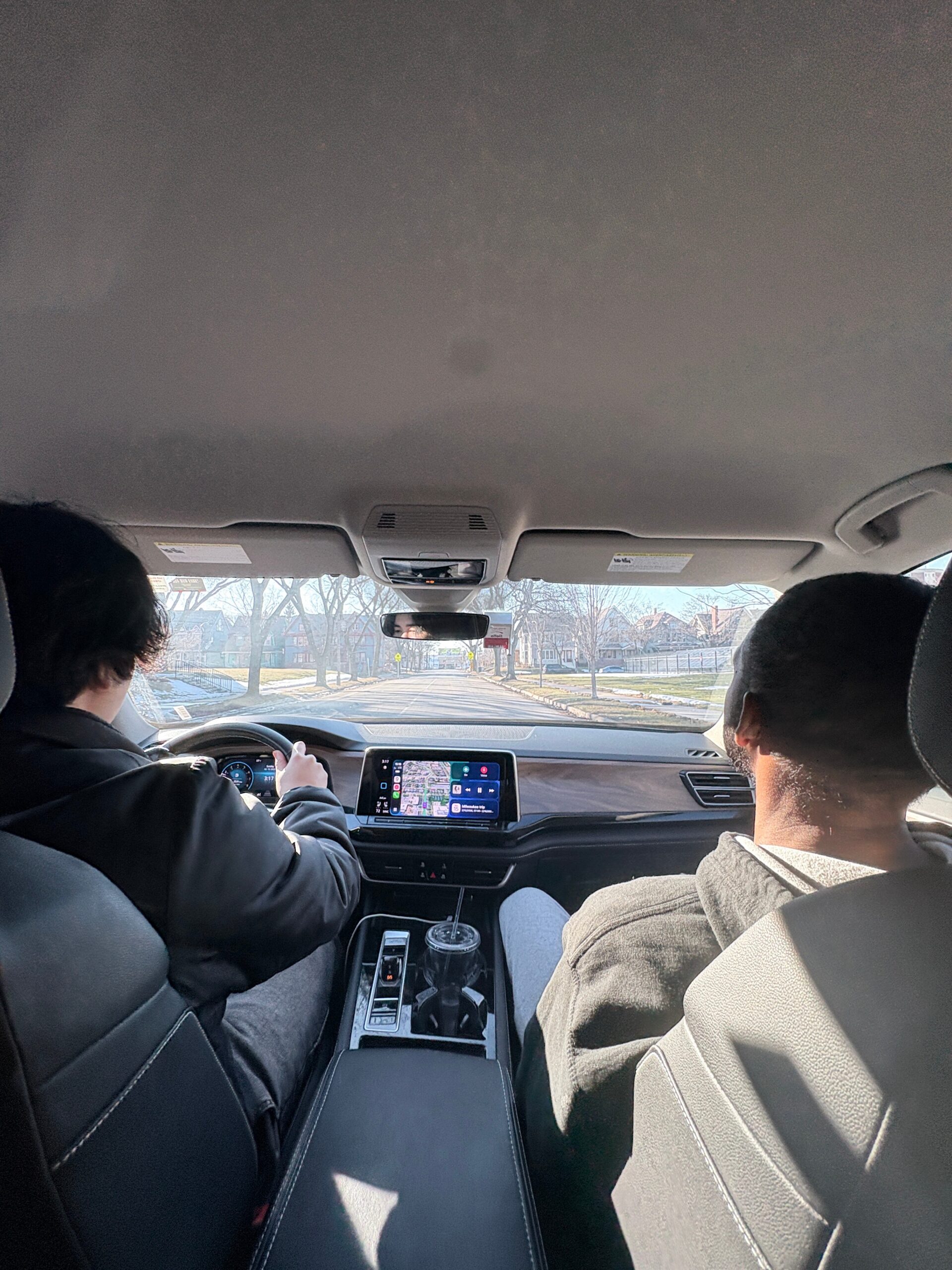 View from the back seat of a car showing the driver on the left and a passenger on the right, with the car's dashboard and a navigation screen visible; outside, a suburban street with bare trees.