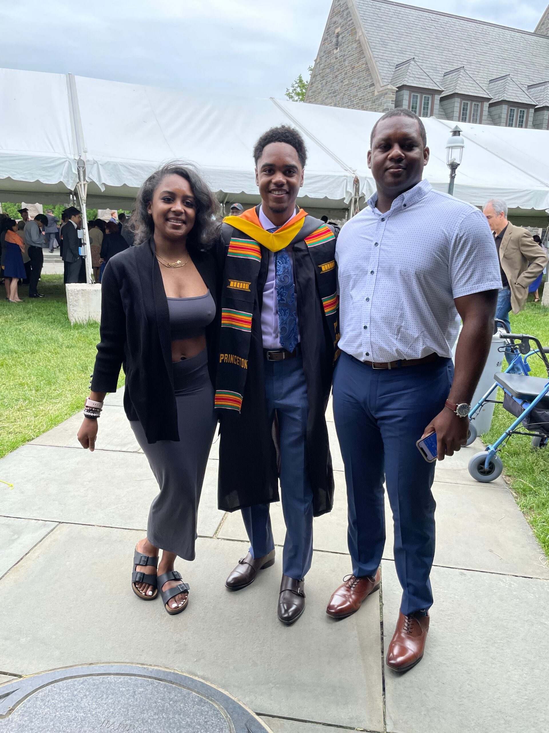 Three people pose together at an outdoor graduation event; the center person wears a black graduation gown with a colorful stole, flanked by a woman on the left and a man on the right, with a large white tent behind them.