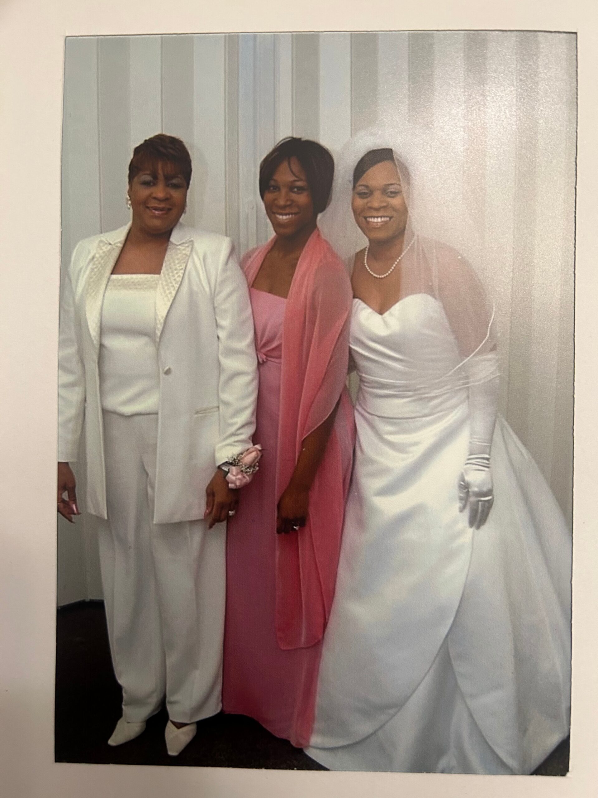 Three women in formal attire pose together: left in a white suit, middle in pink, right in a white wedding gown with veil and pearls