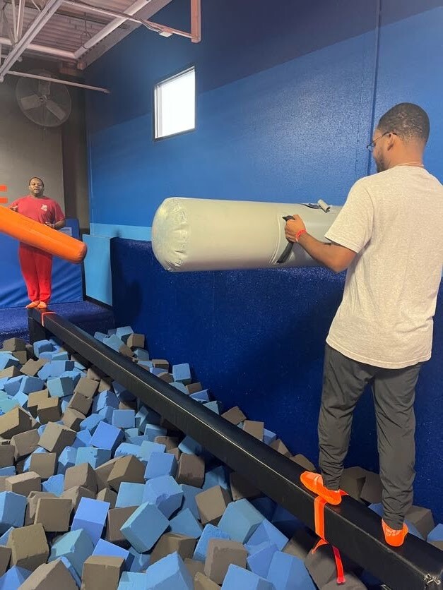Two men balance on a narrow beam above a foam pit in a blue-walled indoor trampoline park, one holding an orange foam prop.