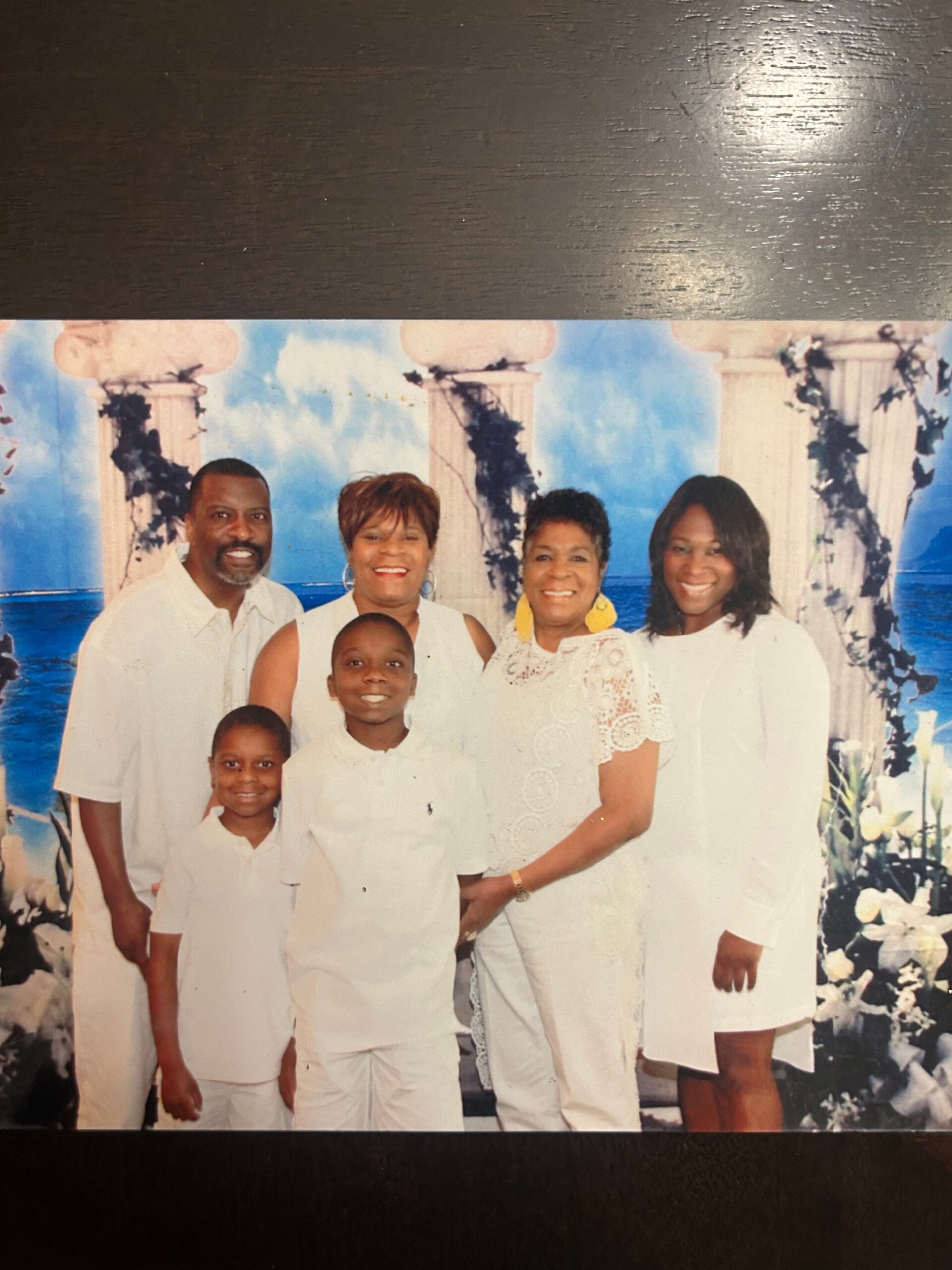 Family group photo outdoors in white outfits, posing in front of a decorative backdrop with columns and blue sky.