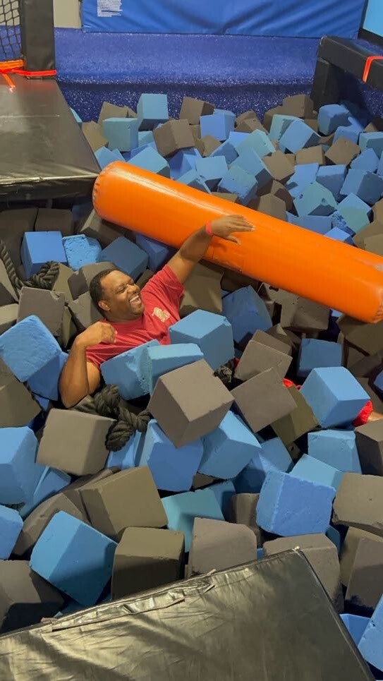 Man in a red shirt laughs while lifting an orange foam log over a pit filled with blue and gray foam cubes at an indoor trampoline park.
