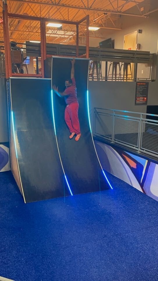 Child in pink clothes climbing a curved indoor play wall with blue neon edges.