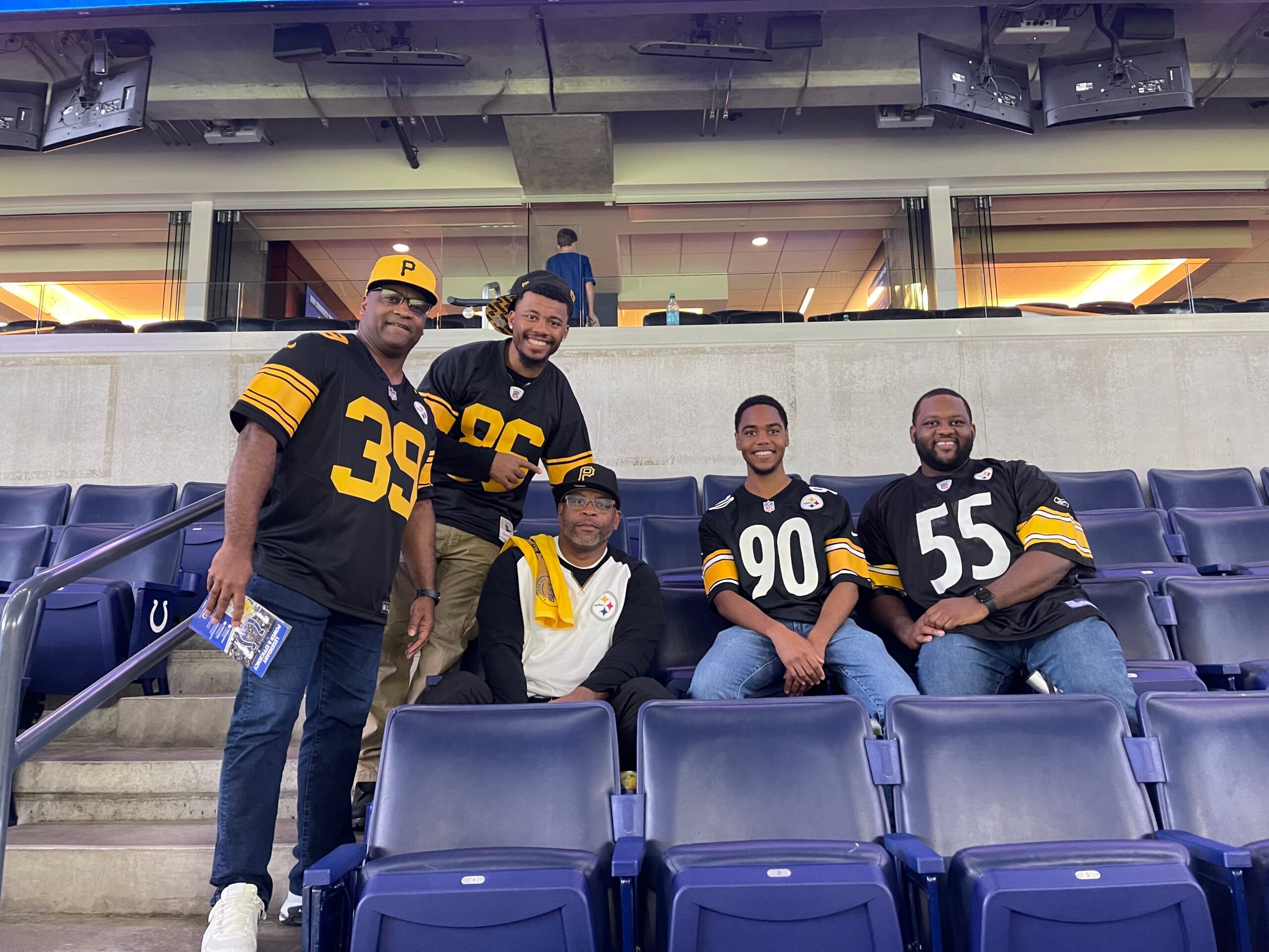 Group of five men in Pittsburgh Steelers jerseys posing in stadium seating, smiling at the camera as fans stand behind them in the stands.