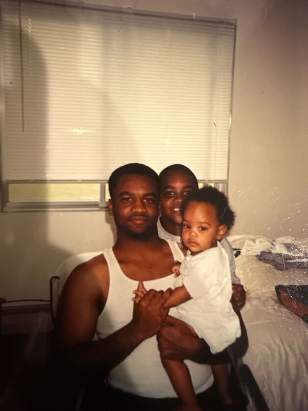 Two men holding a baby in a bedroom, smiling at the camera.