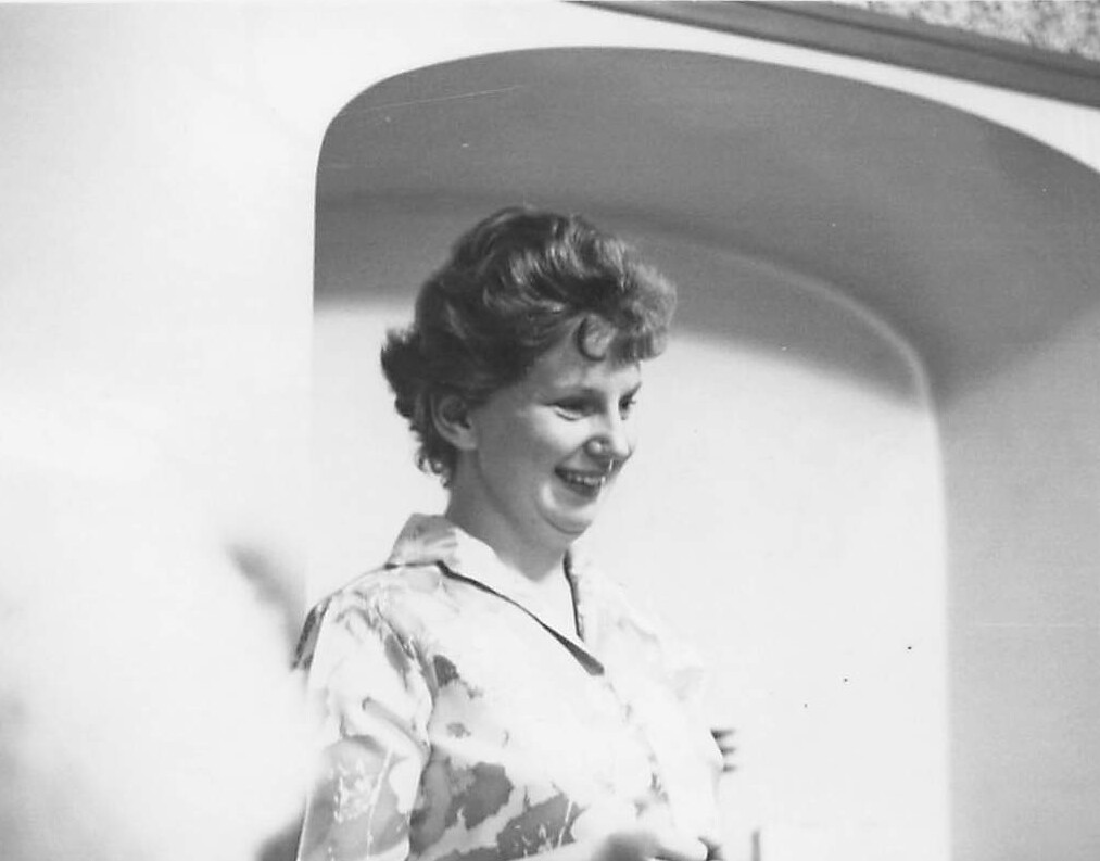 Woman smiling outdoors, standing beside a curved white wall in bright sunlight, wearing a patterned blouse.