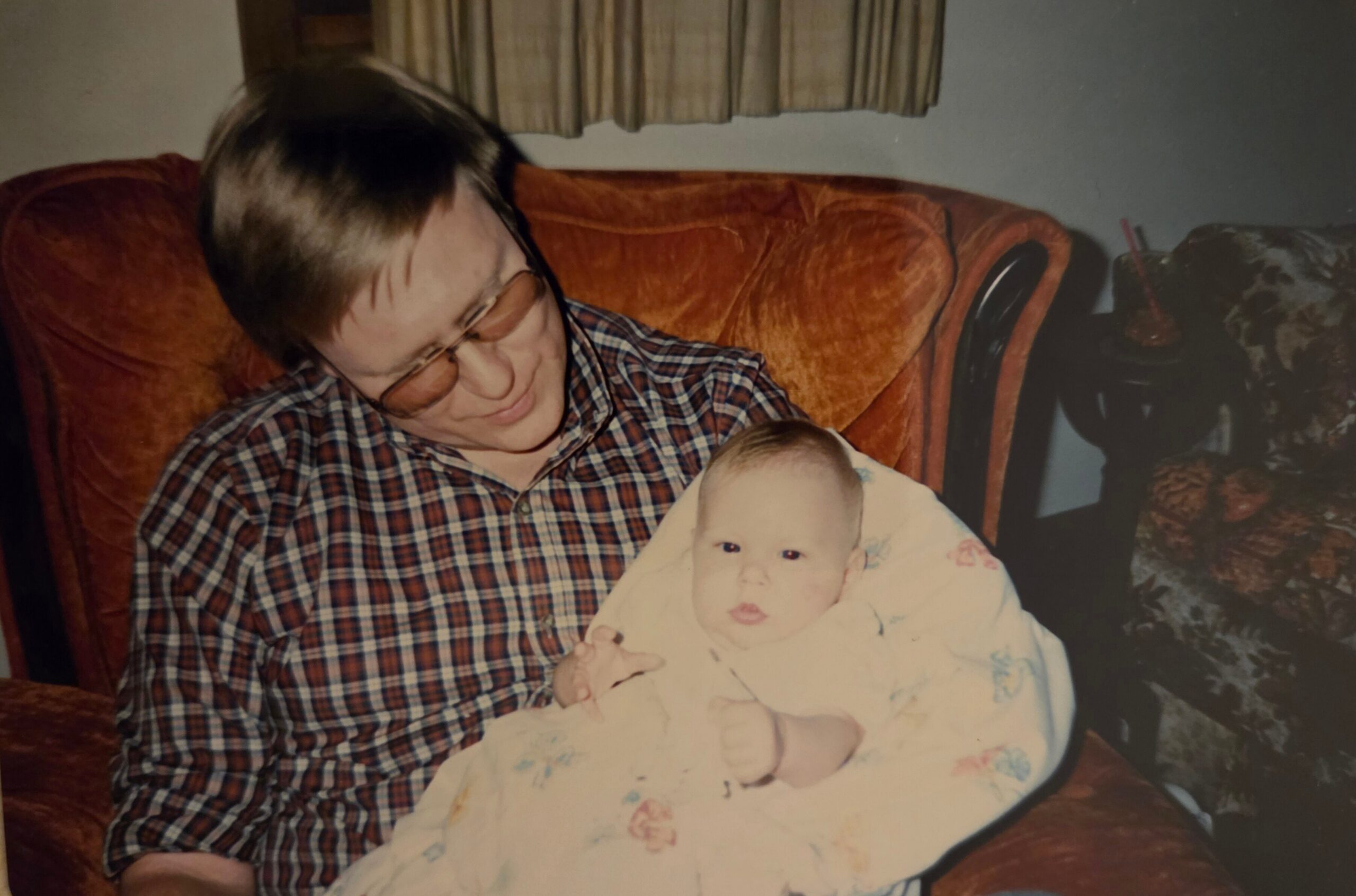 Man in a plaid shirt cradles a swaddled baby on a rust-colored sofa.