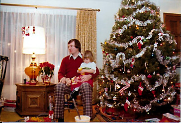 Man sitting with a small child on his lap in a cozy living room, near a decorated Christmas tree with gifts underneath.