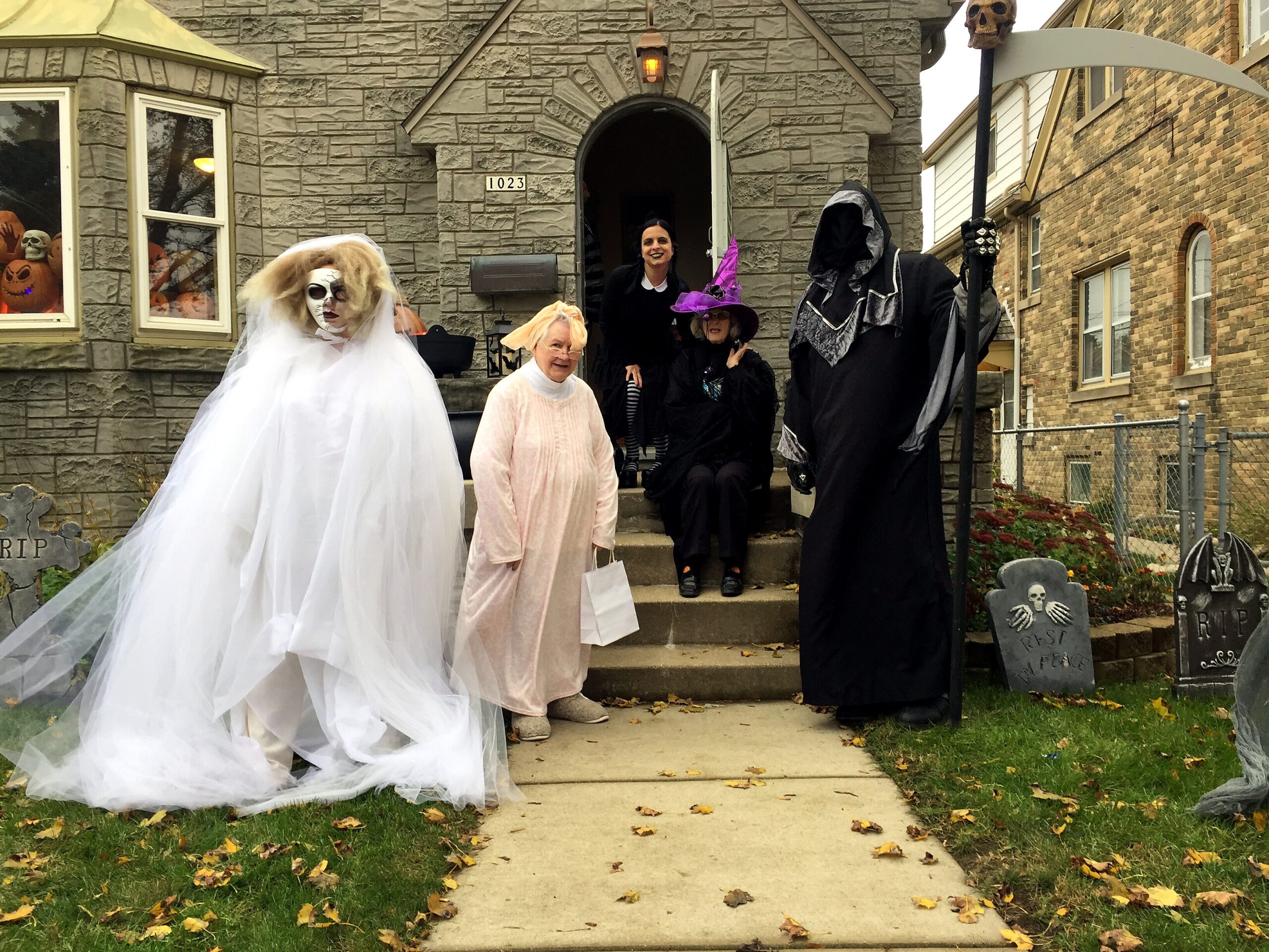Group of people in Halloween costumes on a house steps, including a bride in white, elderly woman, and dark cloaked figures with a scythe and witch hats, with pumpkins in windows and tombstones nearby.