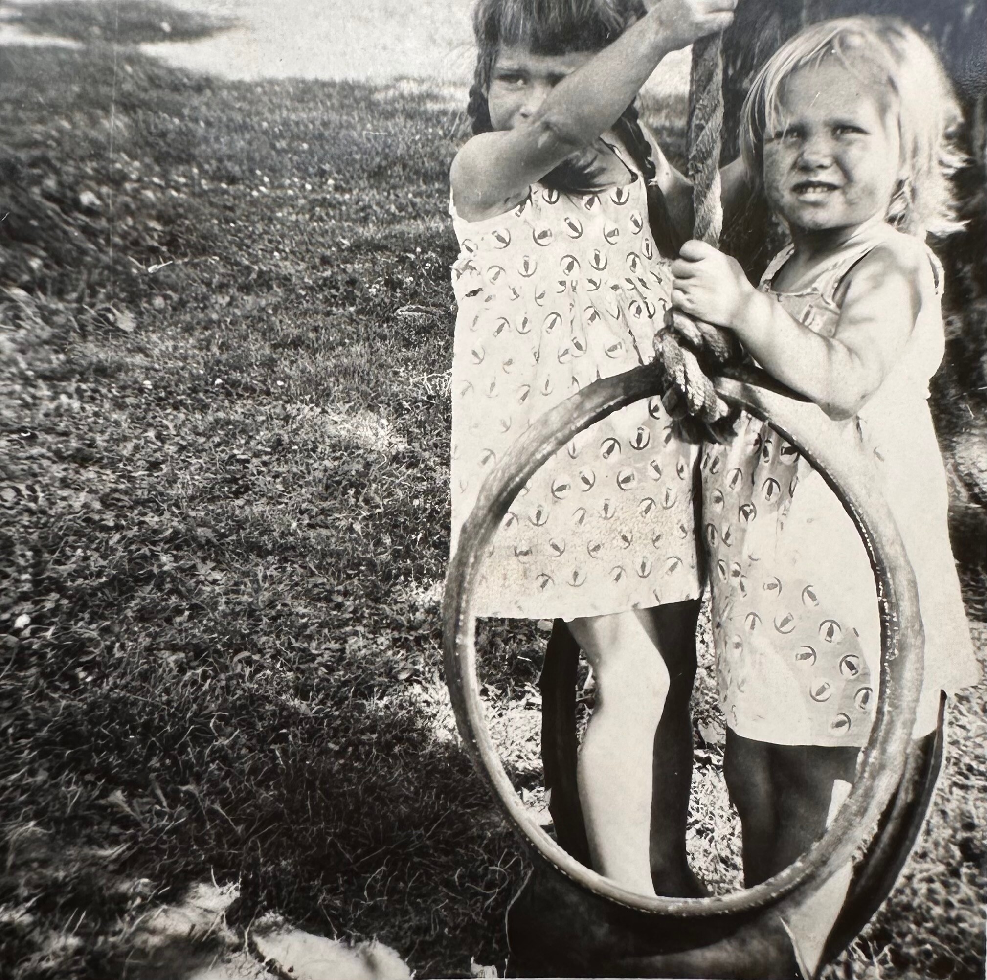 Two young children standing outside by a tree in a grassy field, one shielding their face with an arm and the other looking at the camera.