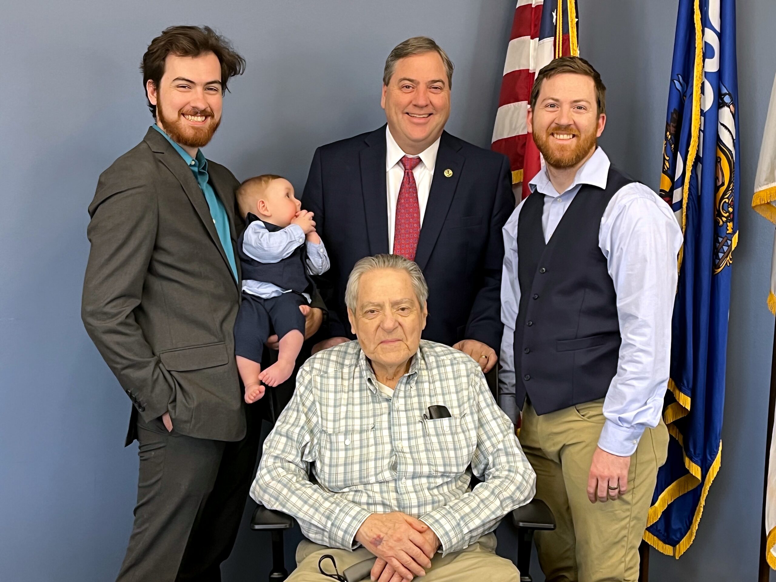 Smiling group photo with an elderly man in a wheelchair at center, flanked by men in suits and a baby being held by one of them, flags in the background.