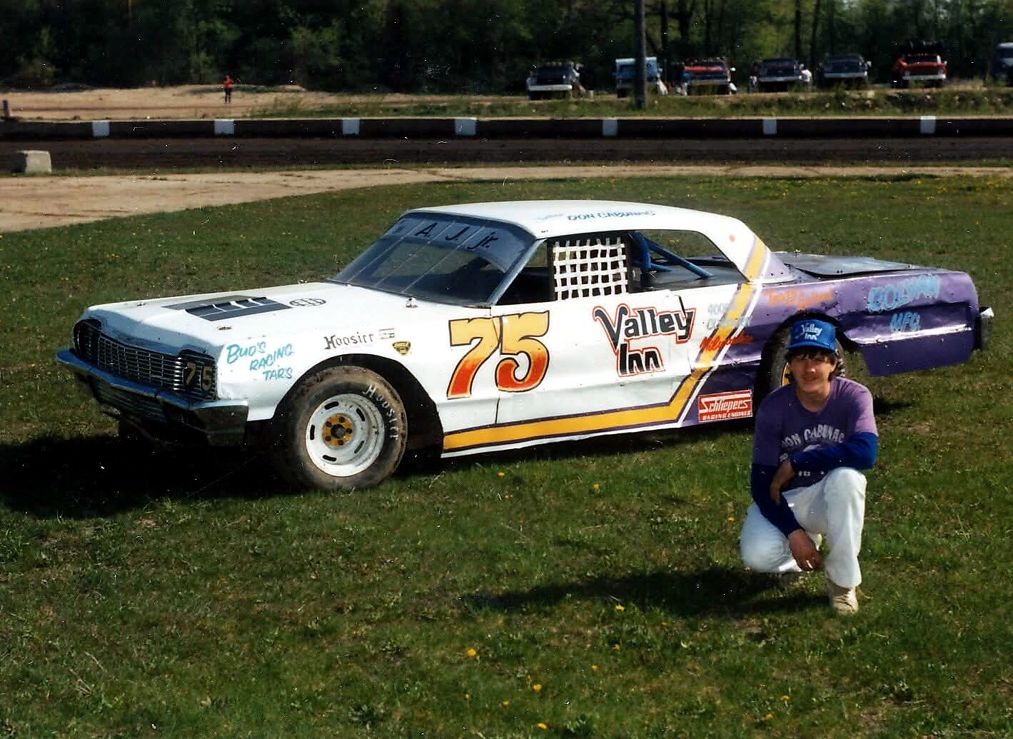 White race car #75 with colorful Valley Inn sponsorship on a grassy field; a person in a blue cap kneels beside it in the foreground.