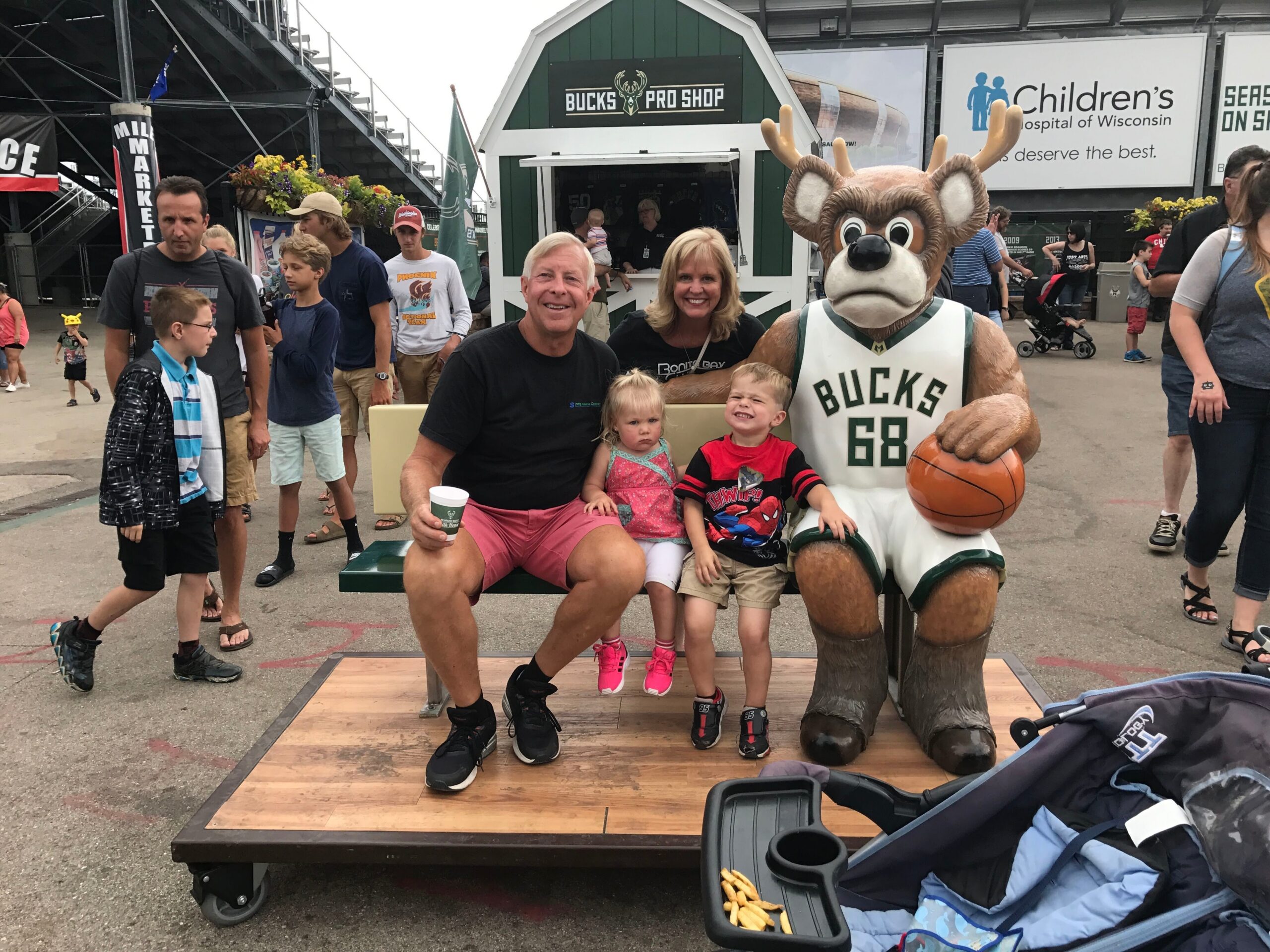 Family seated on a bench with a Bucks basketball mascot at an outdoor fair, posing for a photo with two kids and two adults nearby.