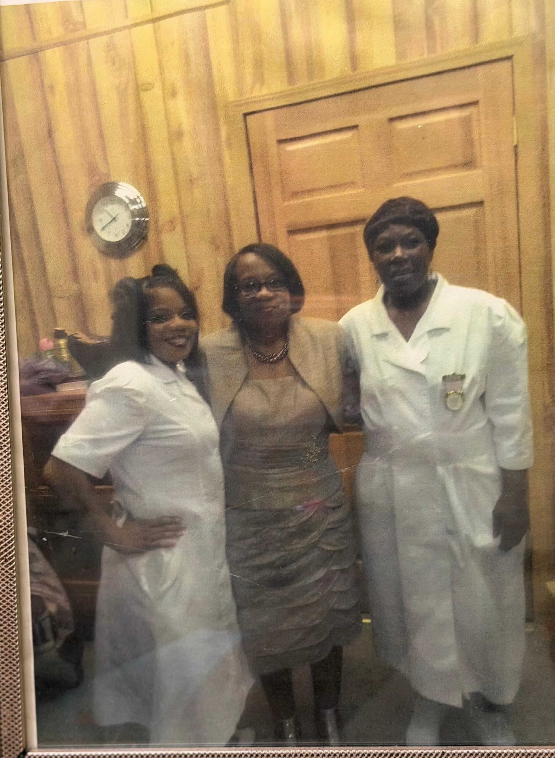 Three adults pose together in a wood-paneled room, with a clock on the wall behind them.