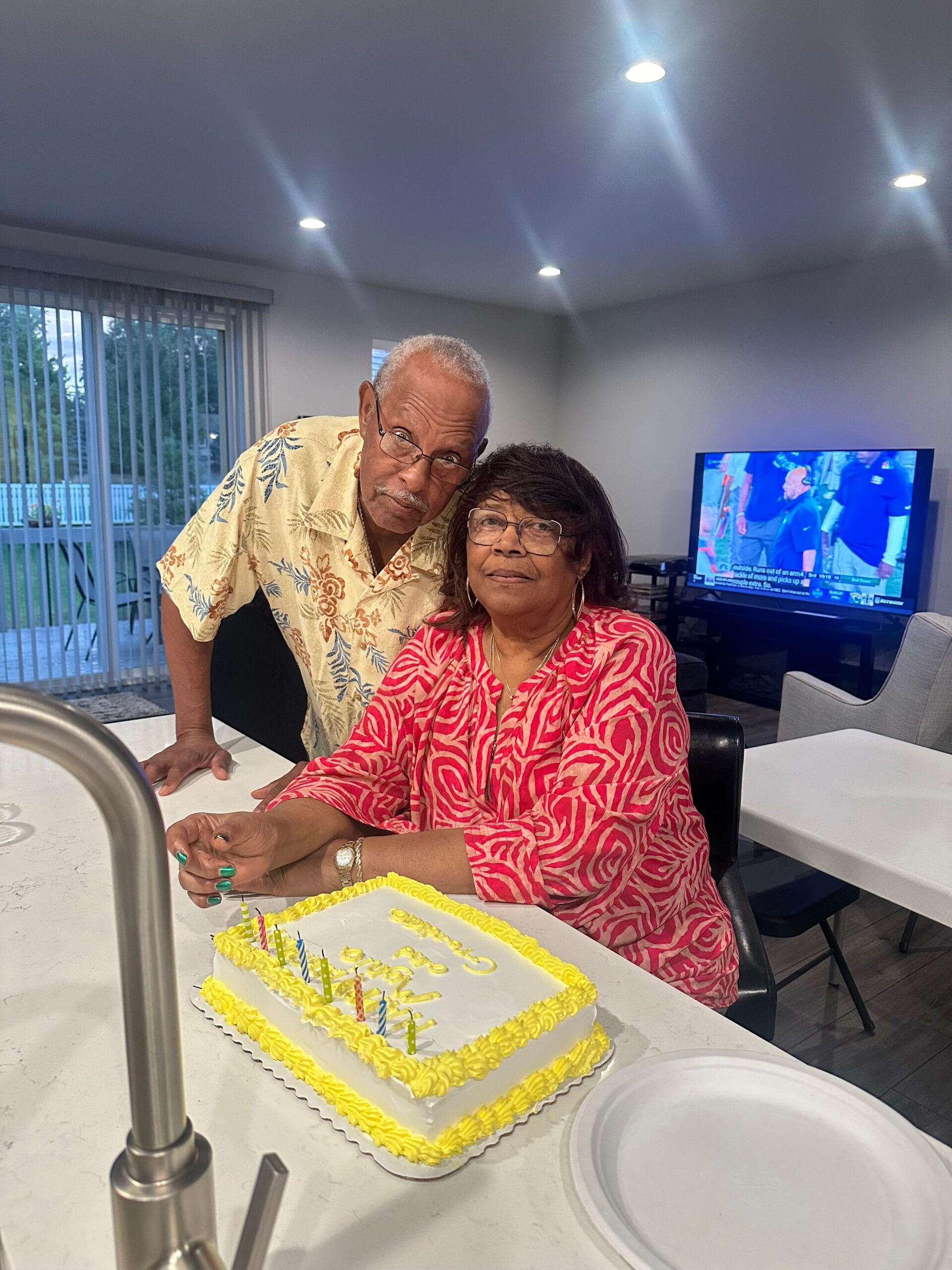 Two elderly adults posing beside a rectangular yellow-iced birthday cake with candles on a kitchen island, woman in a pink patterned top and man in a floral shirt nearby.