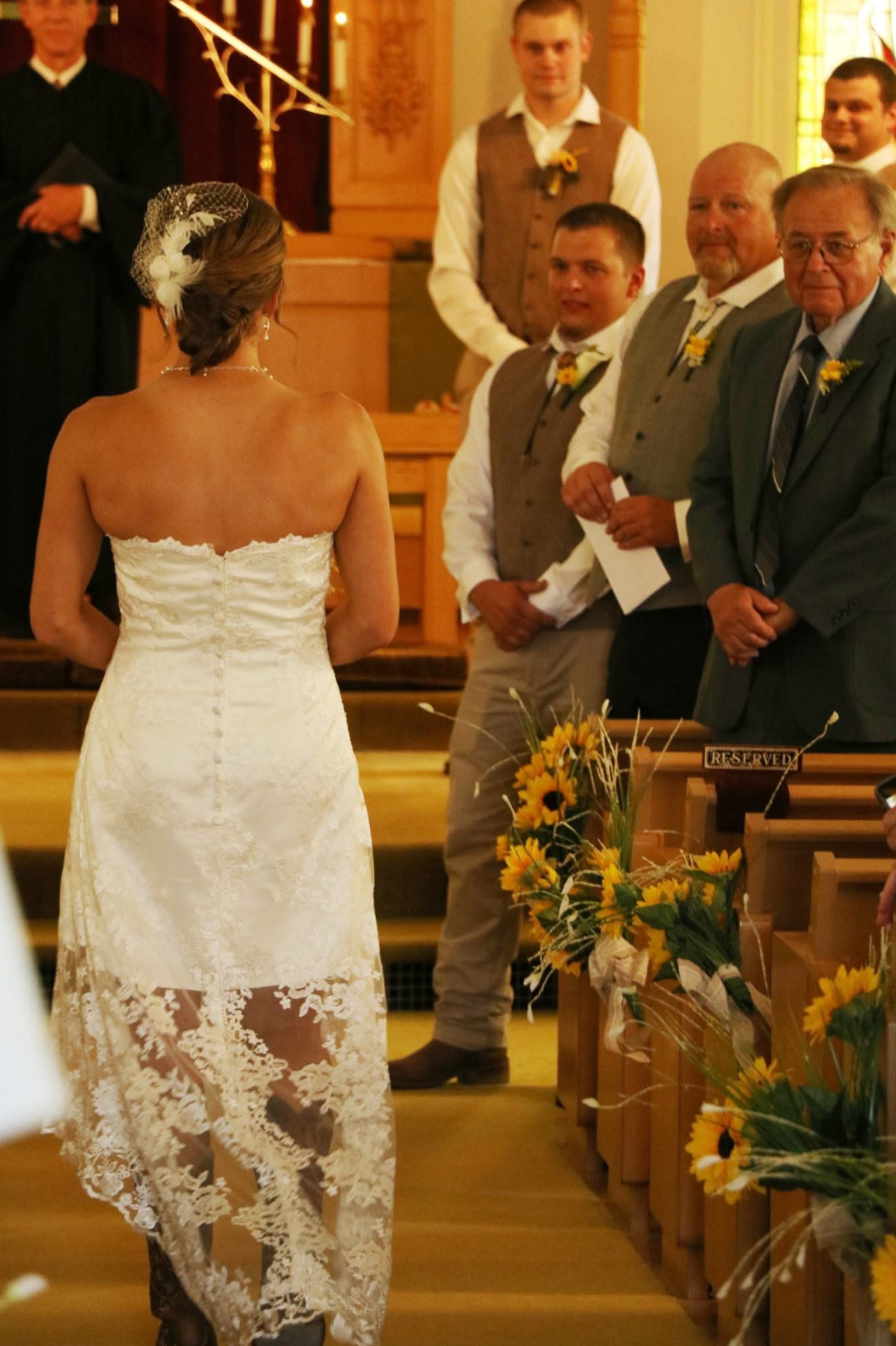 Bride in a white lace strapless gown stands at the altar, facing groomsmen in vests inside a church; sunflower decorations line the pews