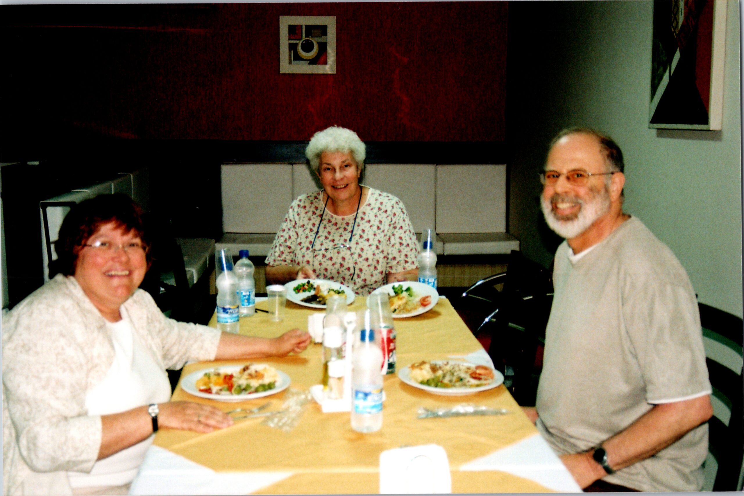 Three seniors sit around a table enjoying a meal together, with plates of food and drinks visible.