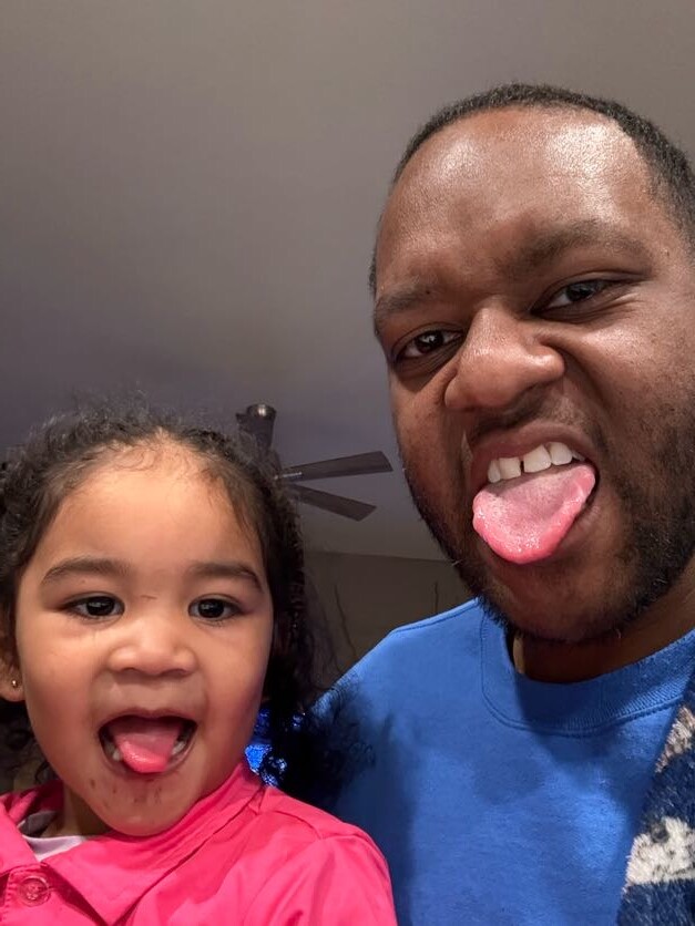 A man and a young girl taking a playful selfie, both sticking out their tongues at the camera.