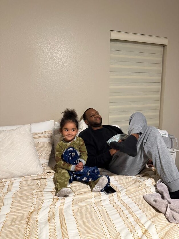 Father and young child sit together on a bed in a cozy bedroom, smiling at the camera.
