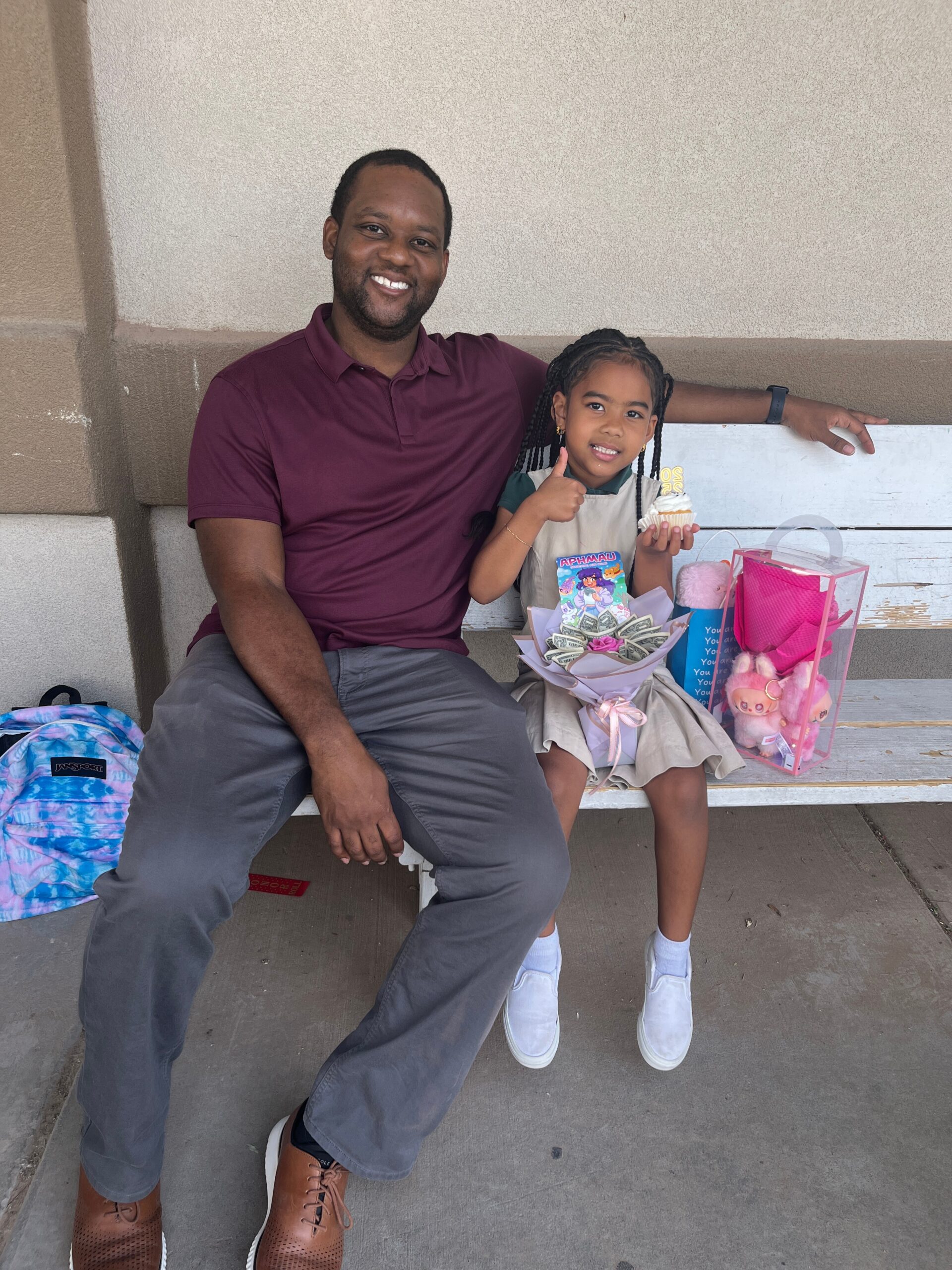 Father and young girl sitting on a bench, smiling, with gift bags and a cupcake nearby.