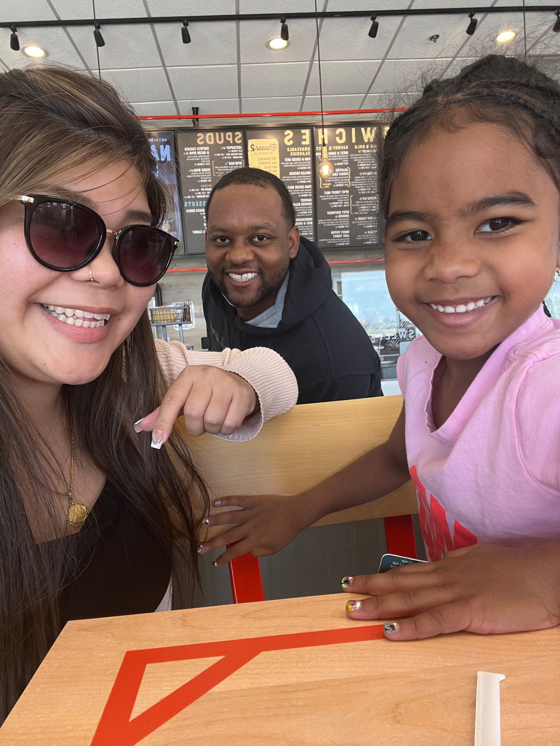 Three people smile at a fast-food restaurant: a woman with sunglasses on the left, a man in the middle, and a girl in pink on the right, with menu boards behind.