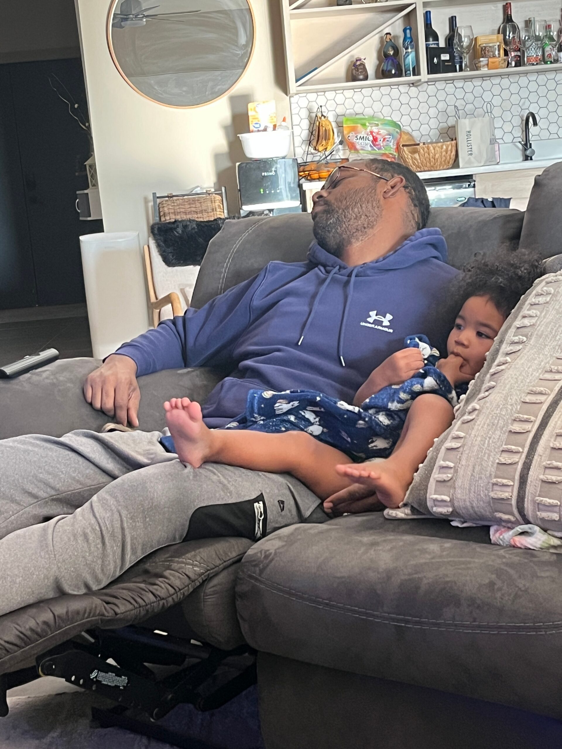 Man in a blue hoodie and glasses reclining on a gray sectional sofa, with a young child snuggled beside him.