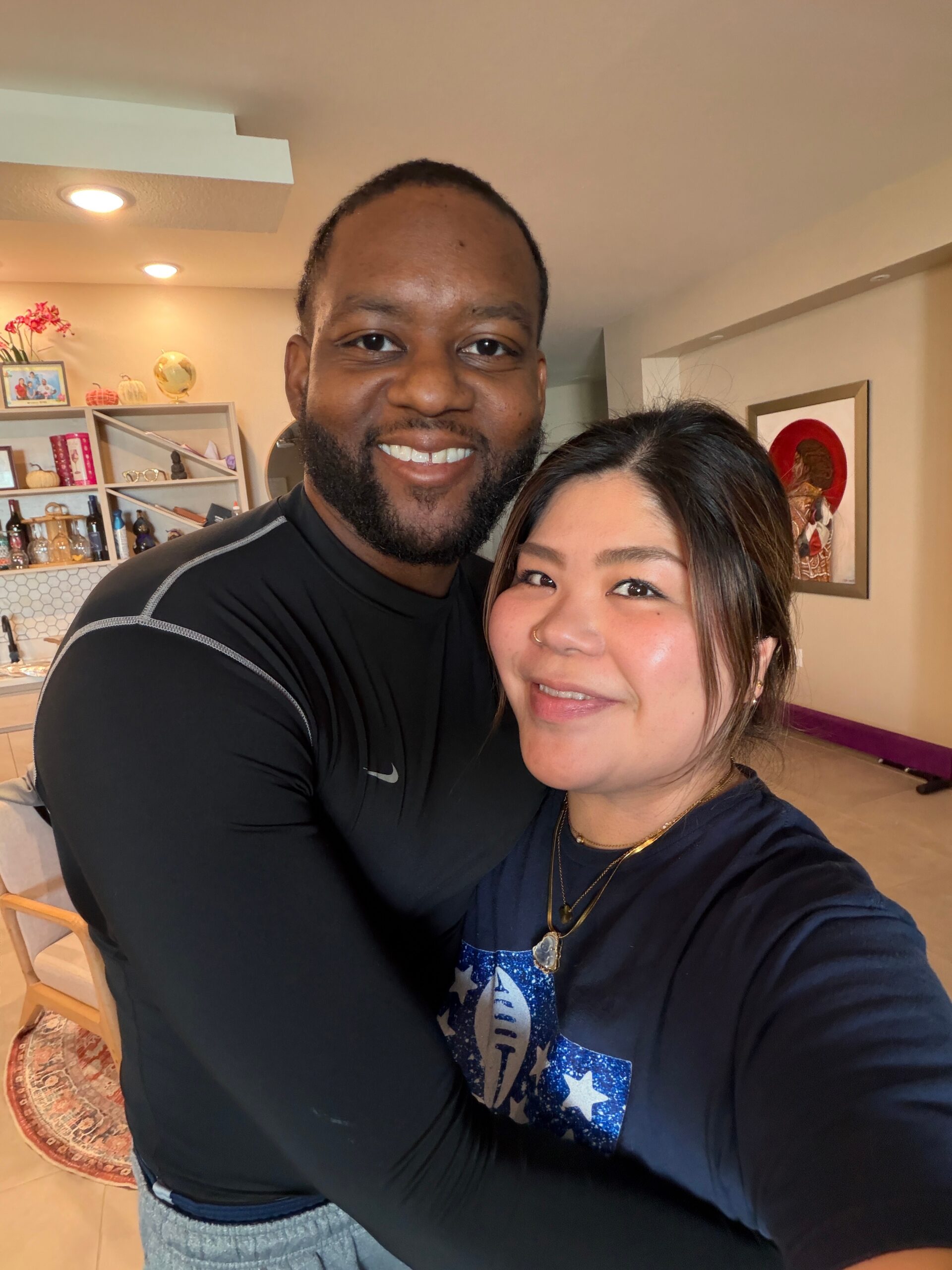 Close-up selfie of a smiling couple indoors, man on the left and woman on the right, both leaning in for the photo in a cozy living area with shelves and decor in the background.
