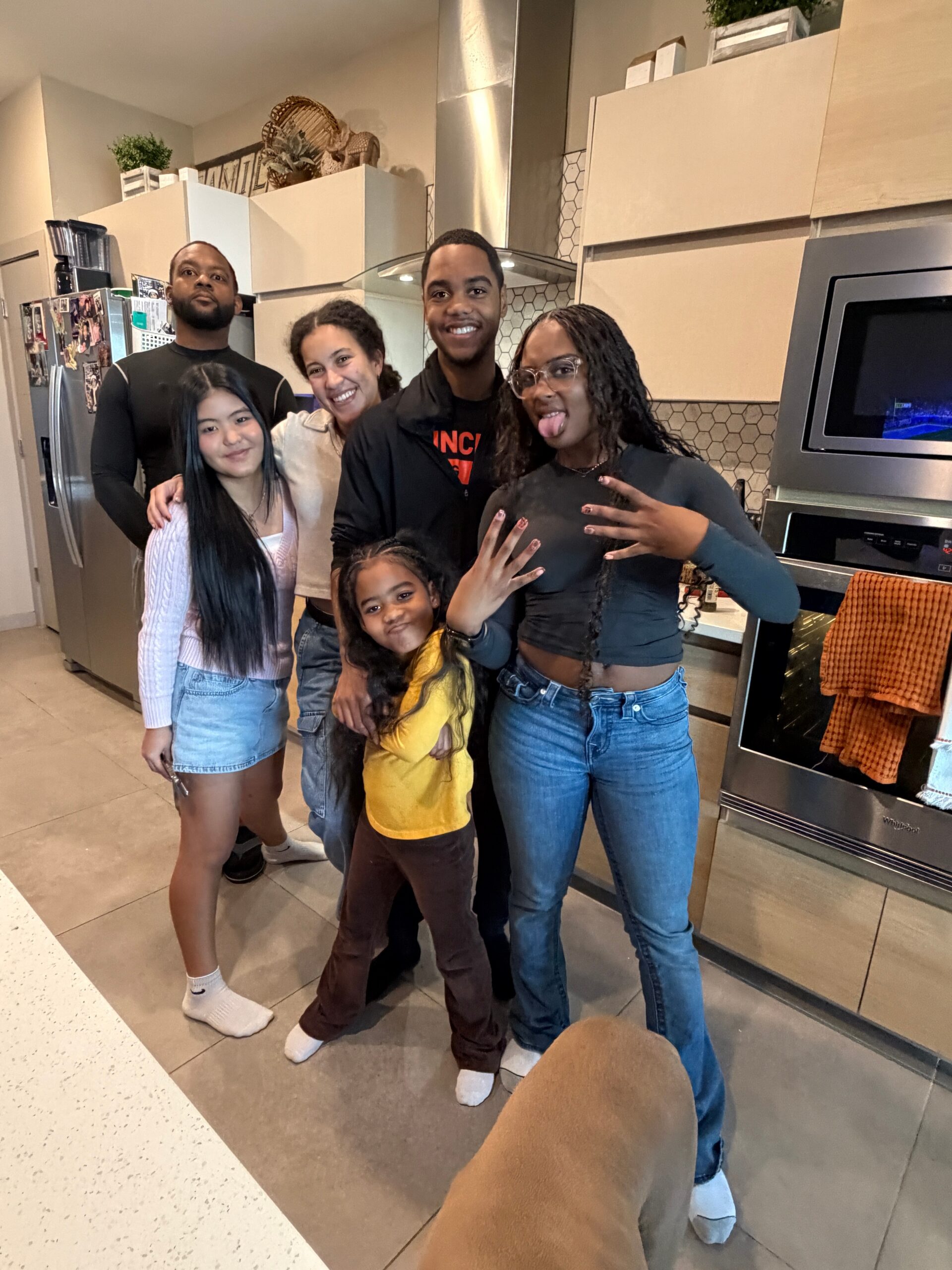 A group of six people posing together in a modern kitchen, smiling and relaxed, with stainless steel appliances and hex tile backsplash in the background.