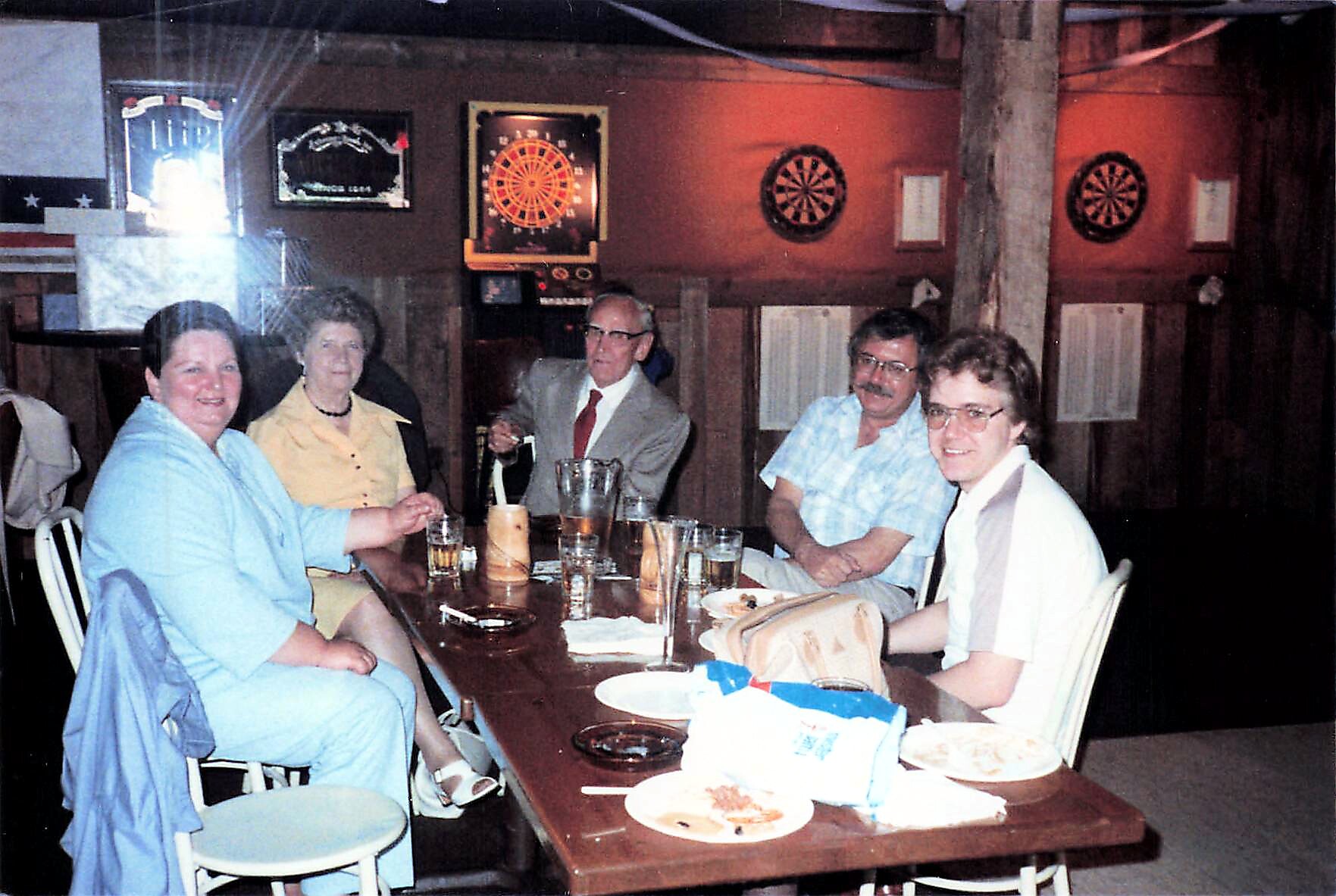 Five adults seated at a long wooden table in a dim bar, with drinks and plates, dartboards on the back wall behind them.