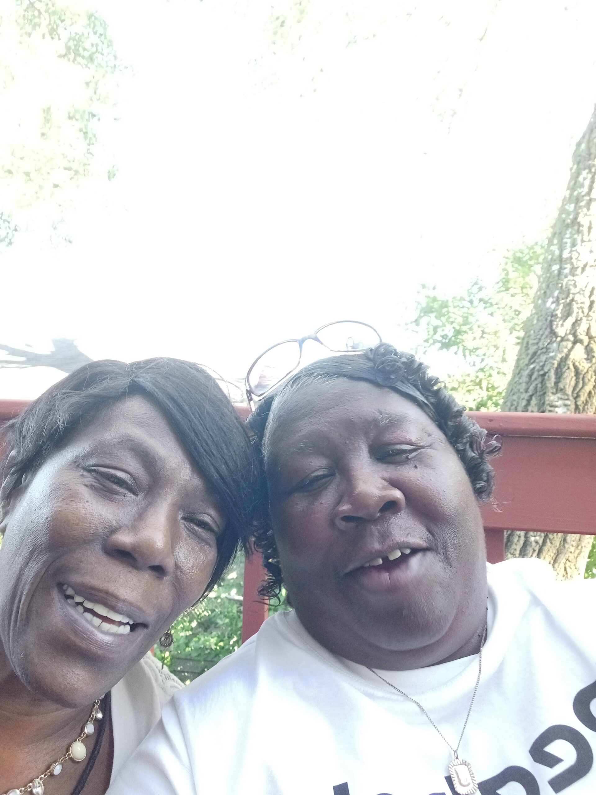 Two women smiling for a selfie outdoors on a sunny day beside a tree and red railing.