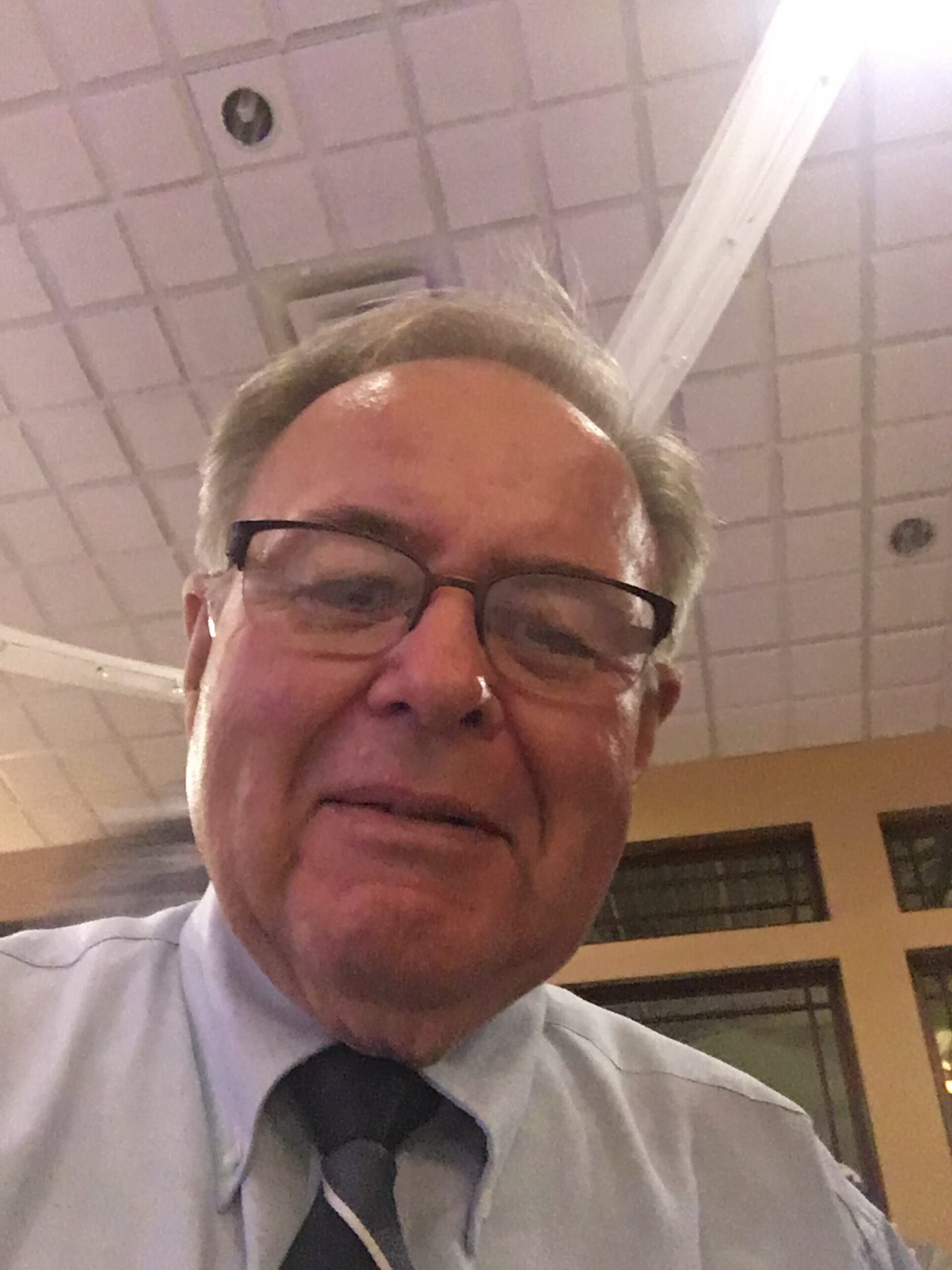 Close-up of a man wearing glasses and a striped tie in an indoor setting, ceiling tiles visible in the background.