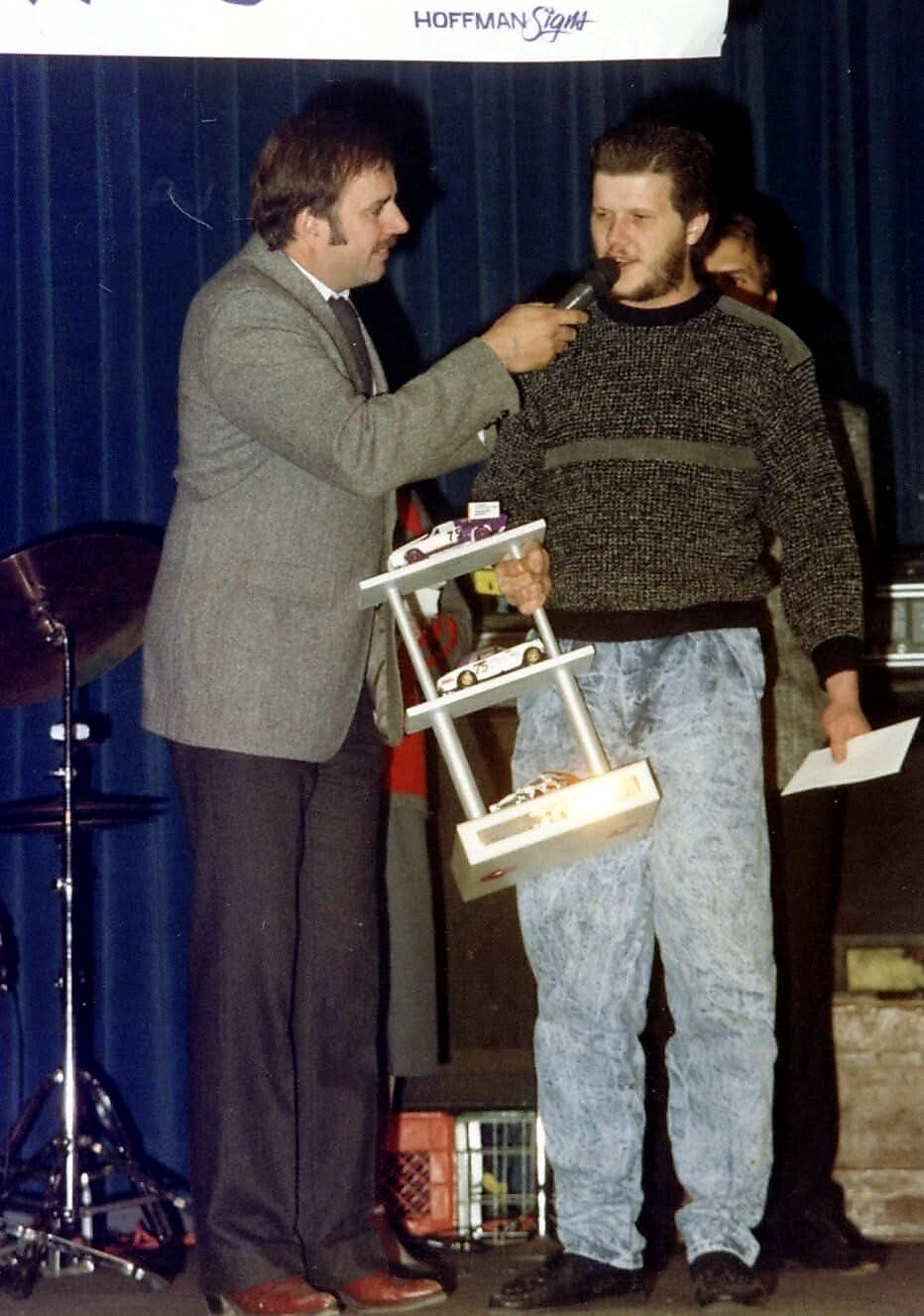 Presenter interviews a man on stage, who holds a small multi-level rack of toy race cars as prizes on a blue-curtain backdrop.