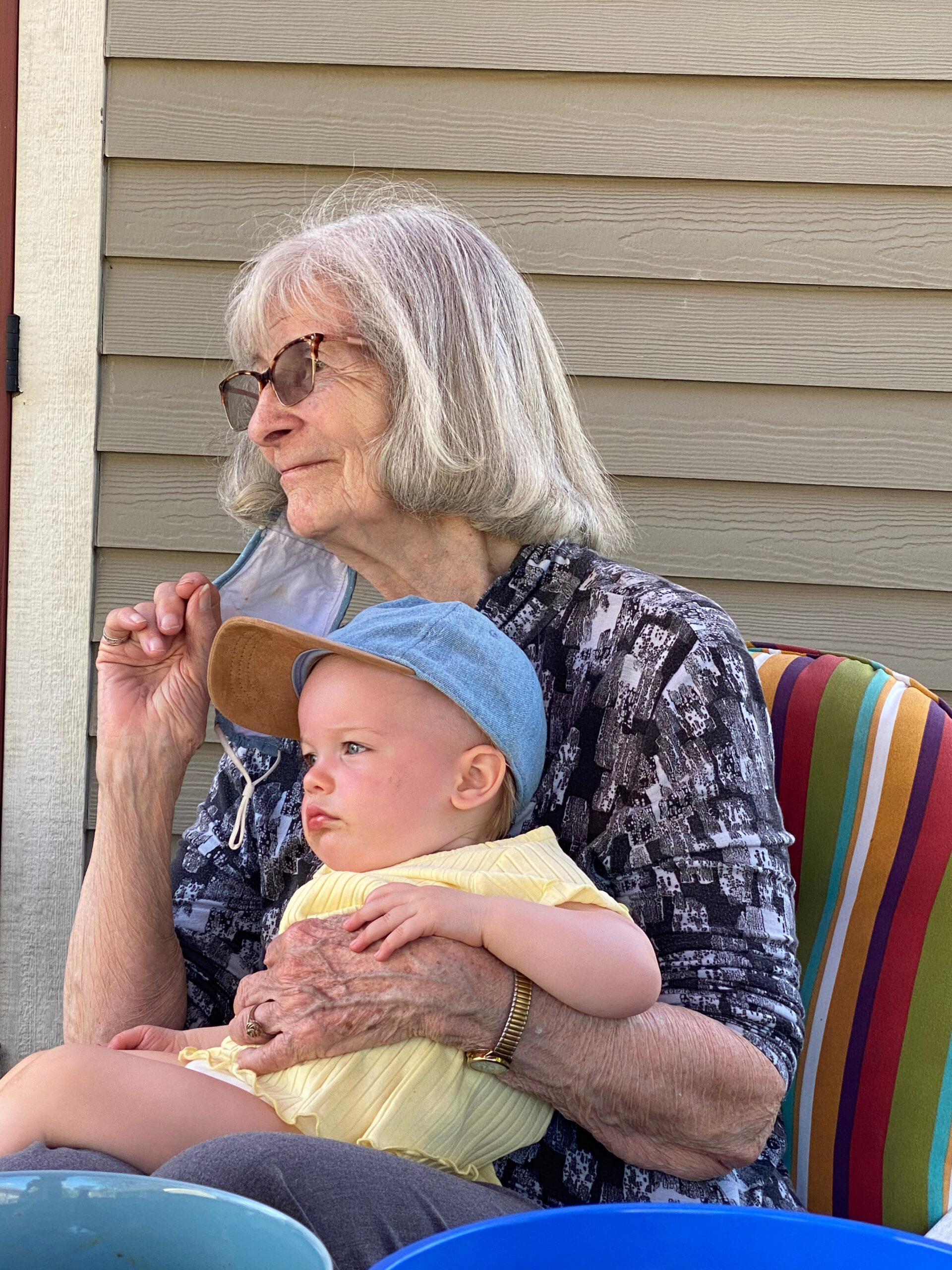 Elderly woman with gray hair and glasses sits outdoors in a striped chair, holding a baby in a yellow outfit and blue hat.