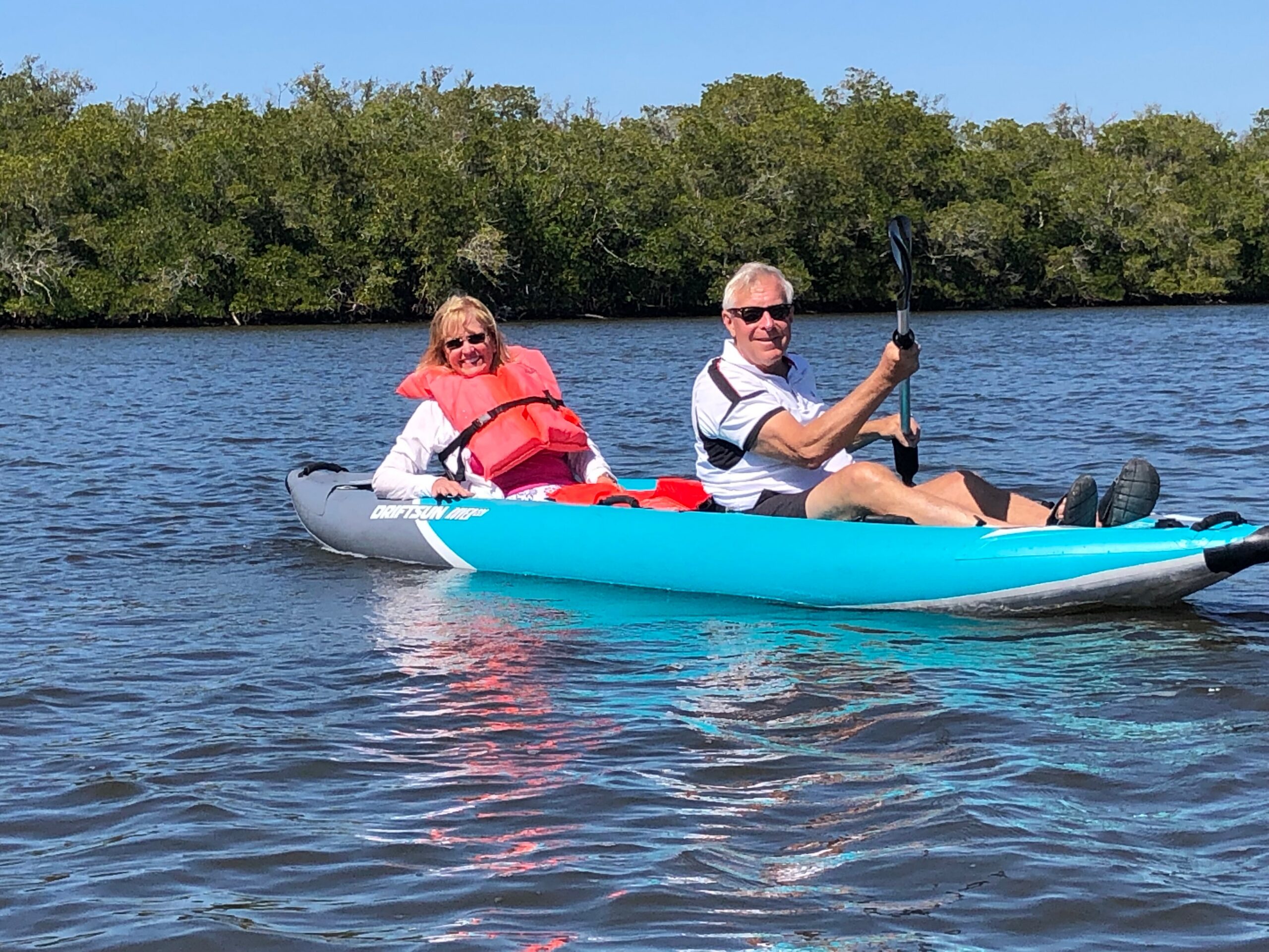 Two people paddling a bright blue tandem kayak on calm water, woman in a pink life jacket and man in sunglasses paddling ahead.