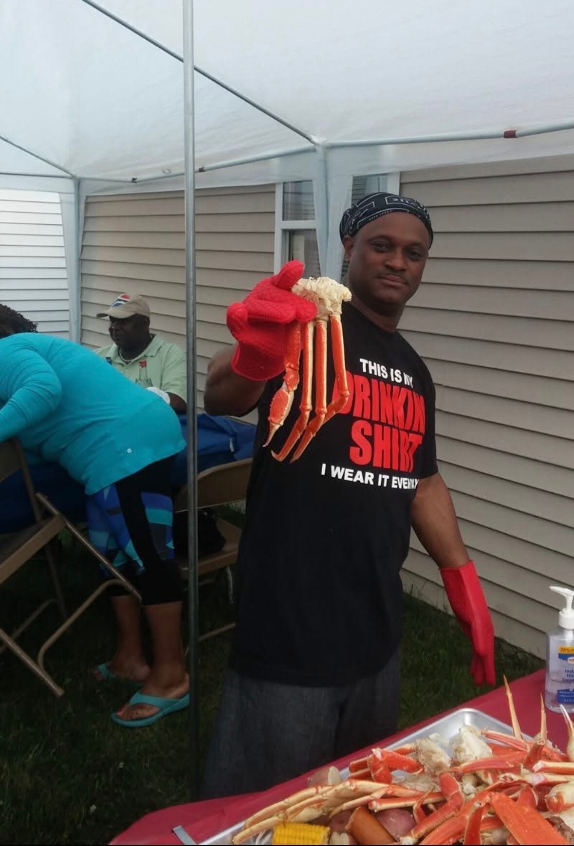 Man in a black shirt and red gloves holds up a cluster of crab legs at a backyard barbecue under a white canopy.