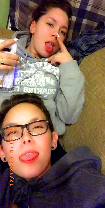 Two friends posing playfully on a couch, both sticking out their tongues; one in a gray hoodie with a finger near the nose, the other wearing glasses and a dark hoodie.