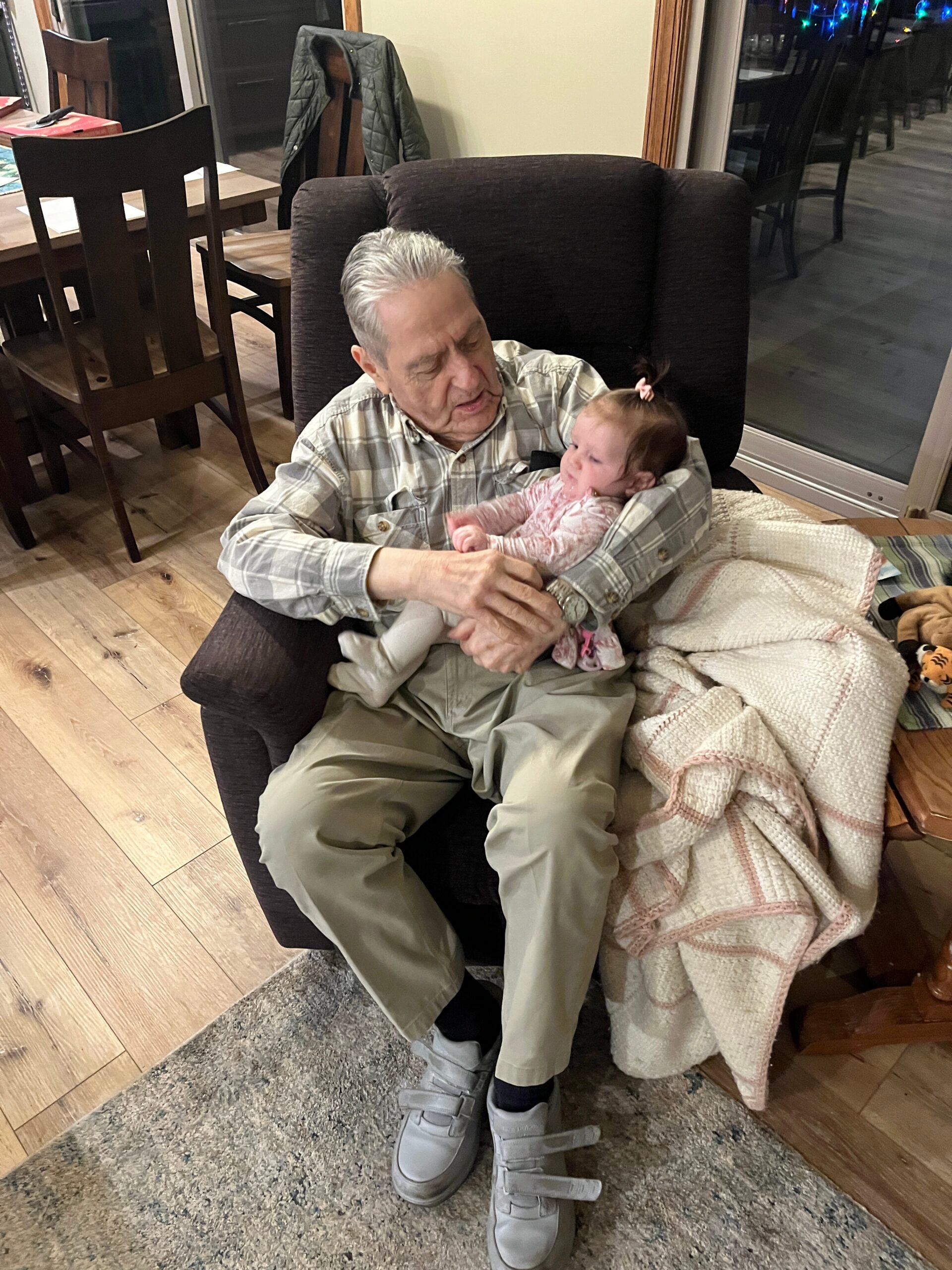 Older man in a plaid shirt sits in a chair, cradling a baby girl in a pink onesie. They are indoors with a blanket nearby and a coat hanging on a chair.